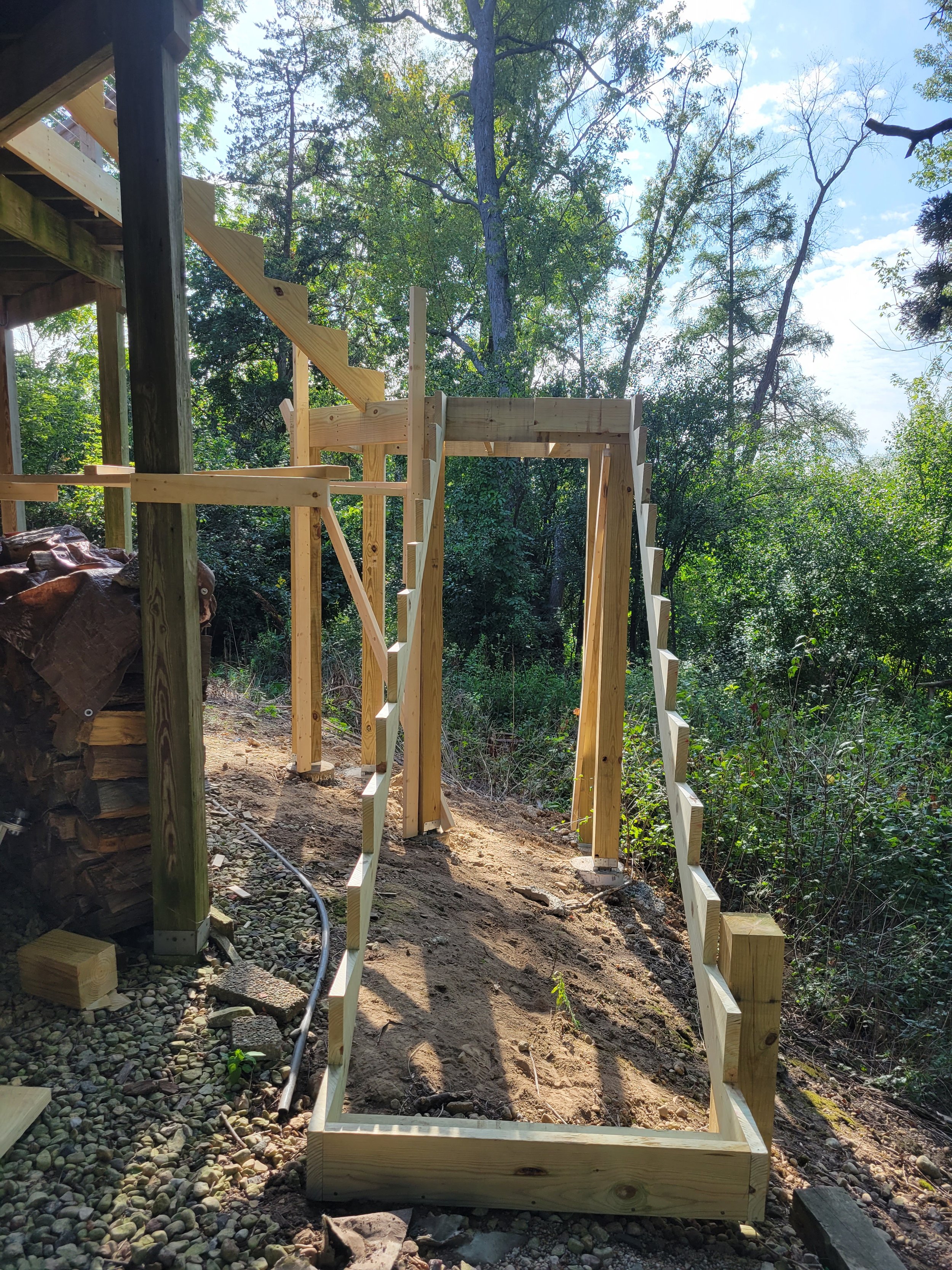 Wooden staircase under construction on a dirt pathway surrounded by trees and greenery, with a pile of wood stacked under the staircase on the left.