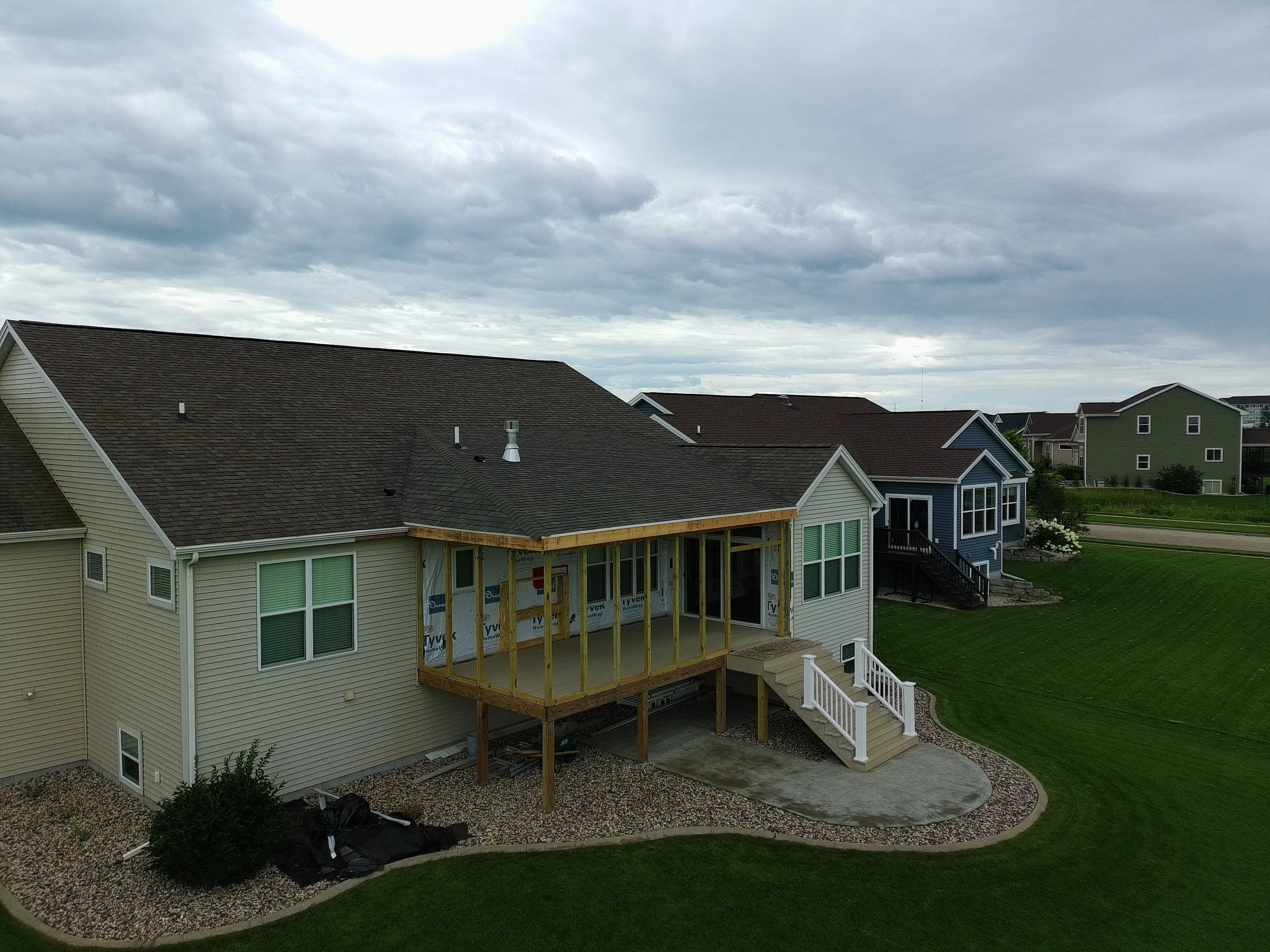 A two-story house under construction with a new wooden deck being built at the back, stairs leading up to it, surrounded by a lawn and neighboring houses under cloudy skies.