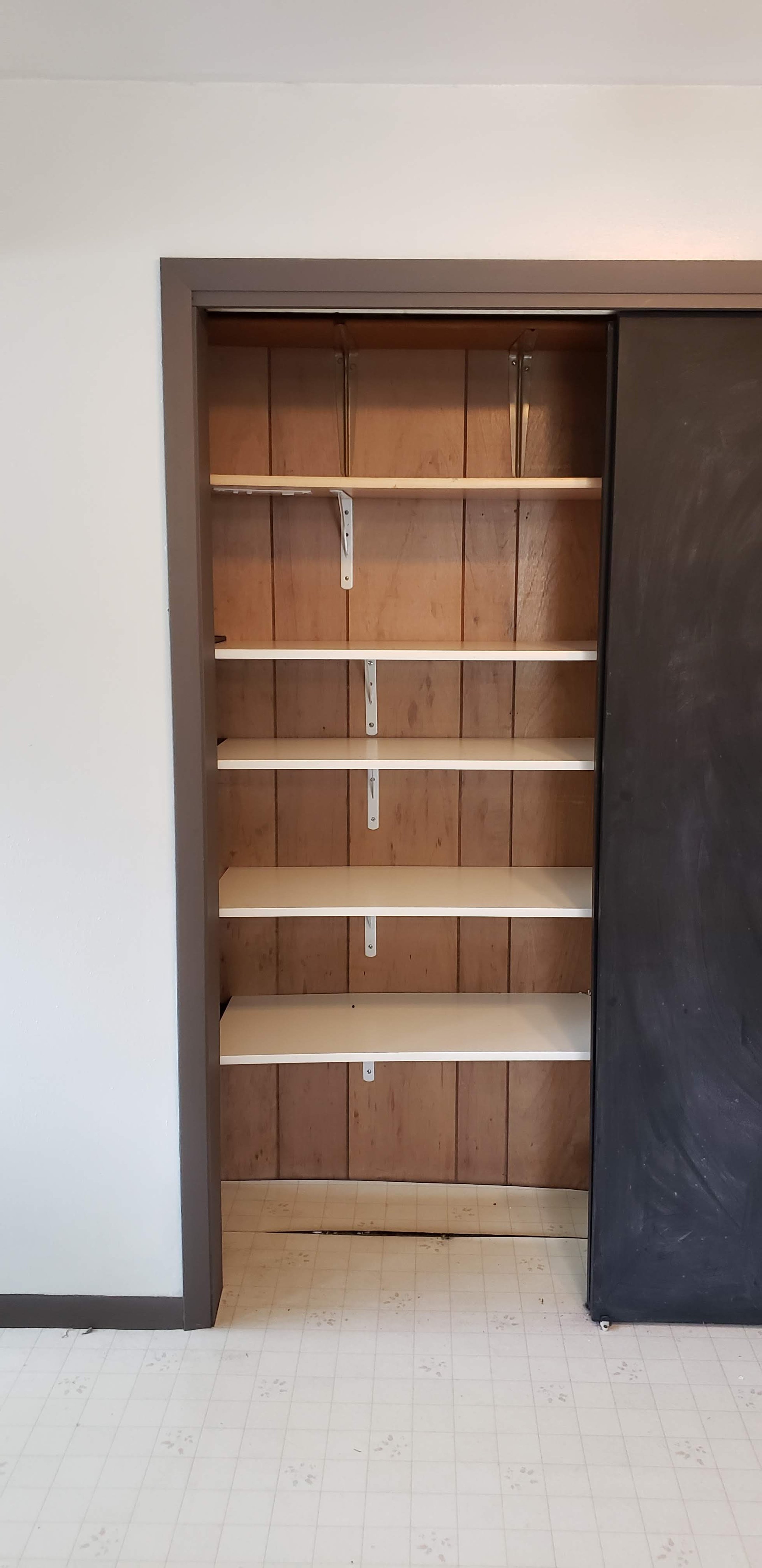 Empty closet with five shelves, partially enclosed with a sliding door, against a brown wood-paneled wall, on a white floor with small square tiles.