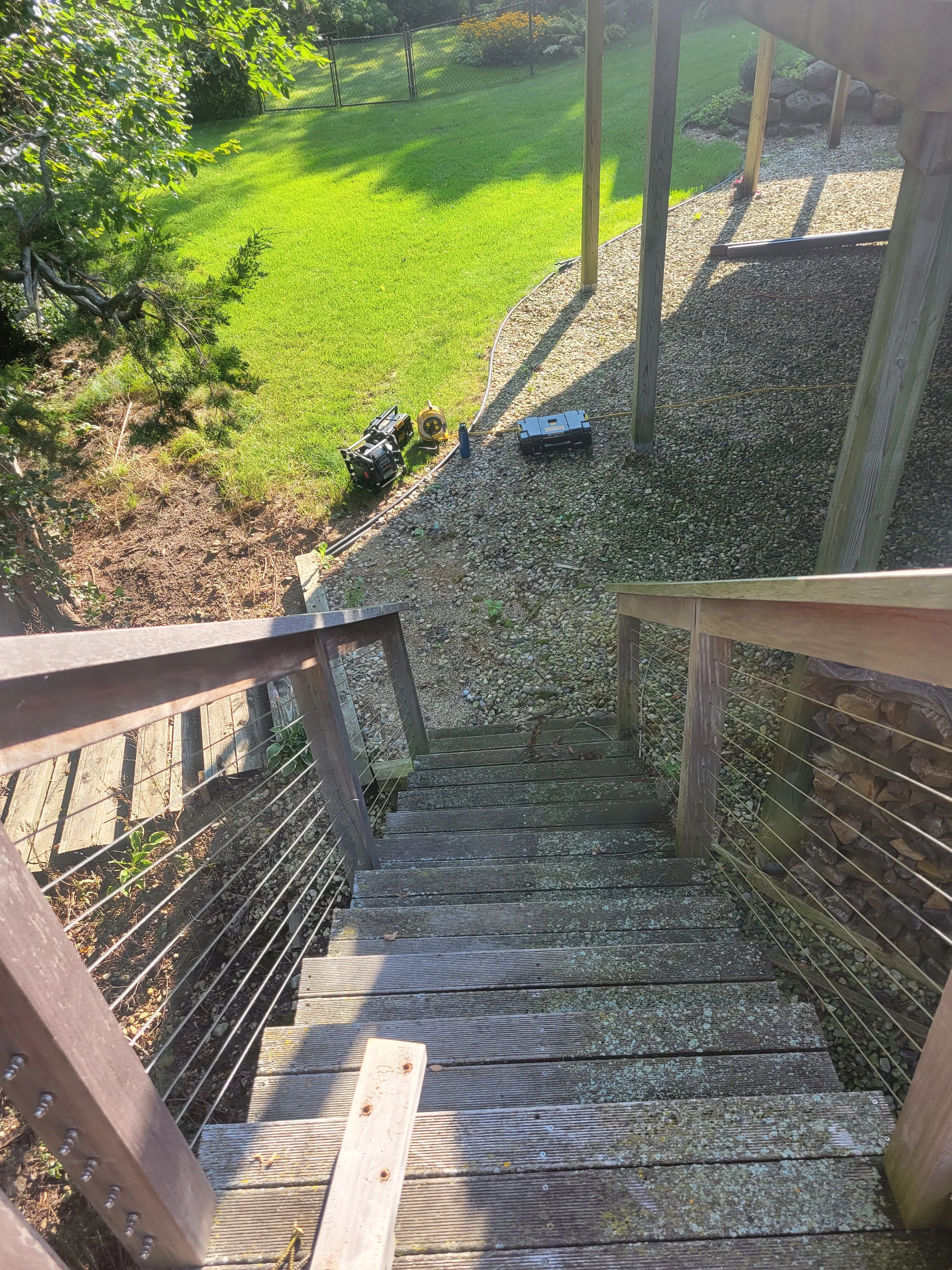 View from top of outdoor wooden staircase looking down to a gravel yard with tools and equipment, surrounded by green grass and trees.