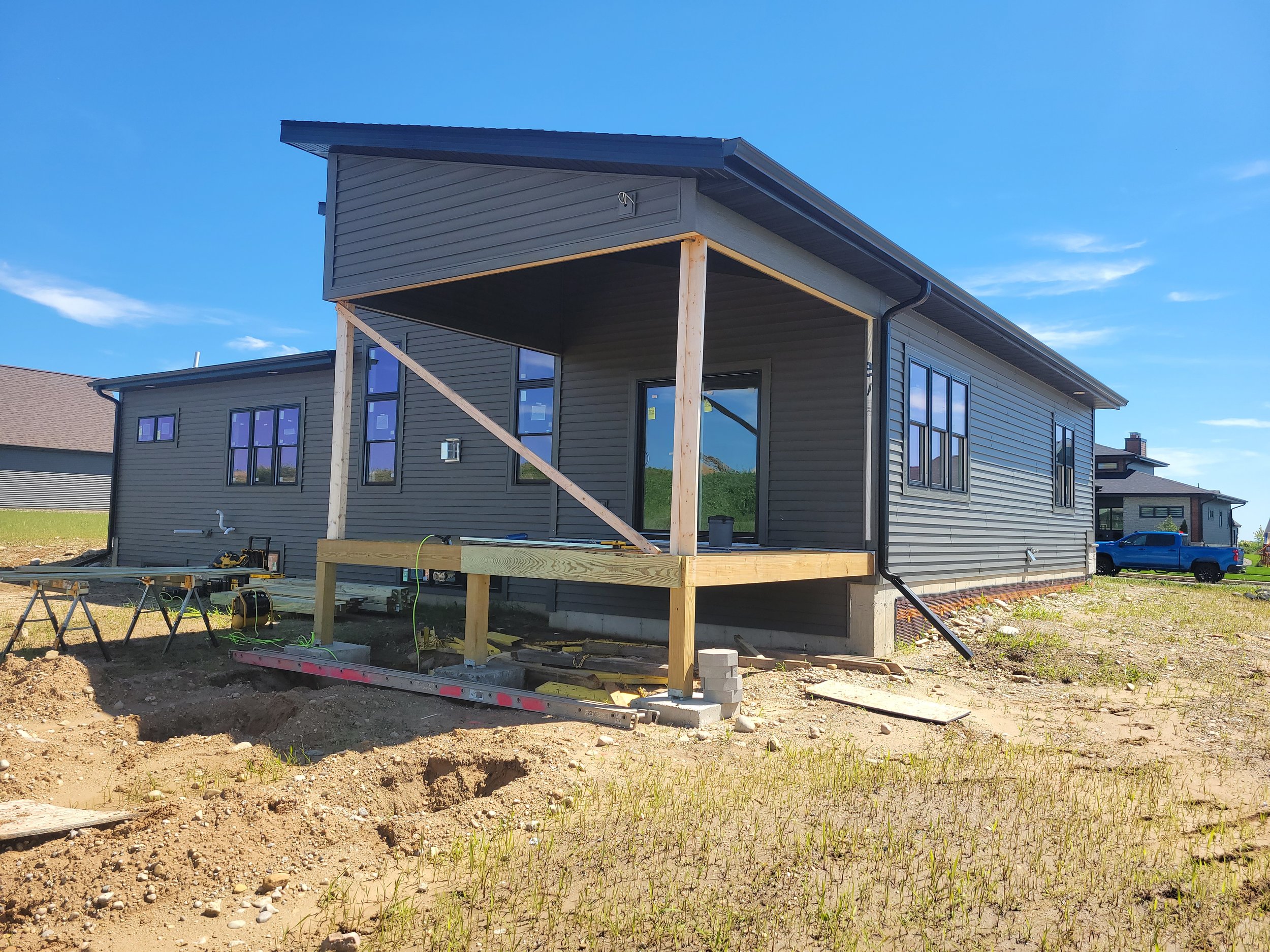 A house under construction with new wooden porch support beams, siding installed, and construction tools on the ground outdoors.