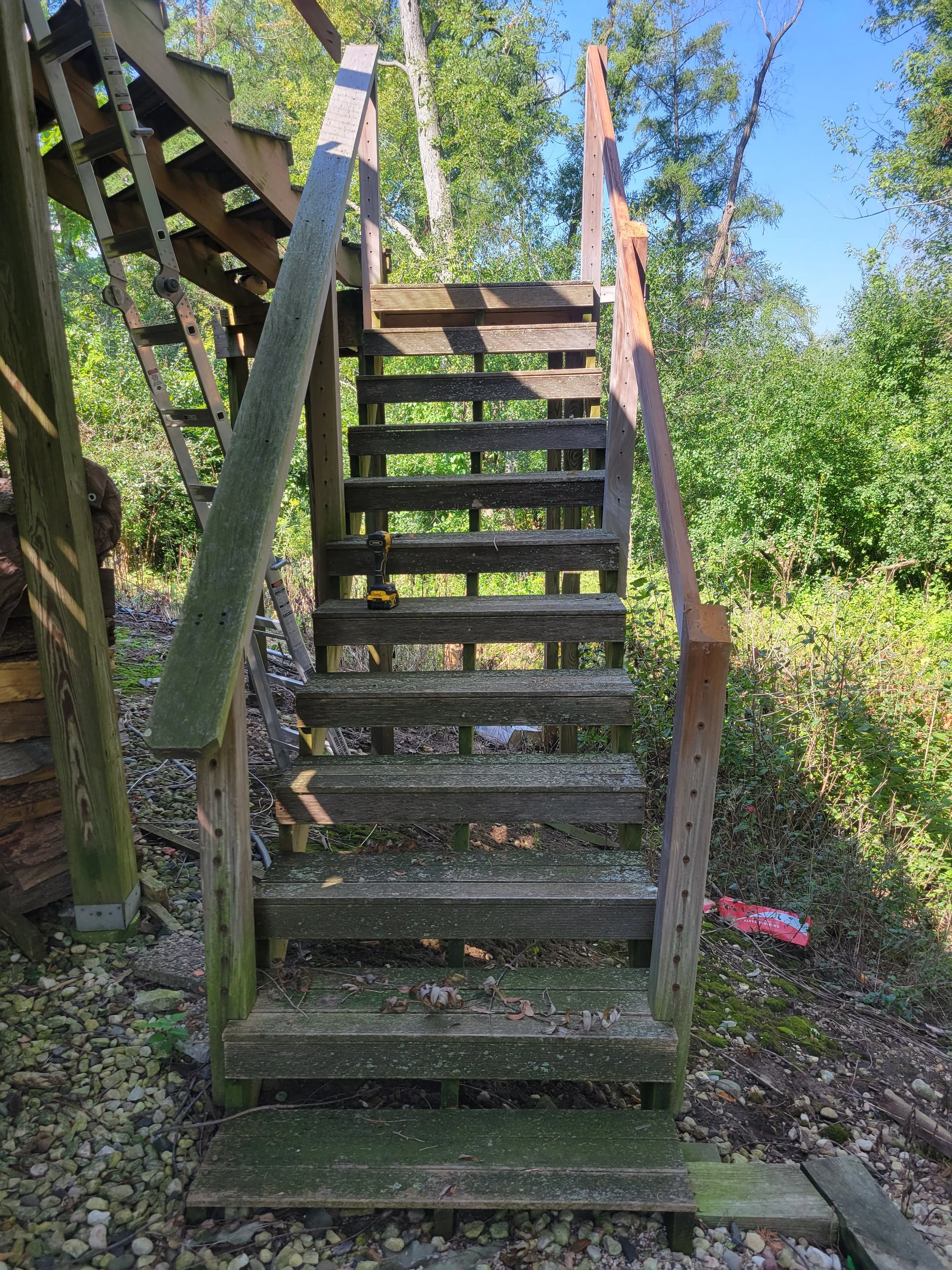 Wooden outdoor staircase under construction in a lush green backyard, with a yellow drill placed on one of the steps, surrounded by trees and foliage.