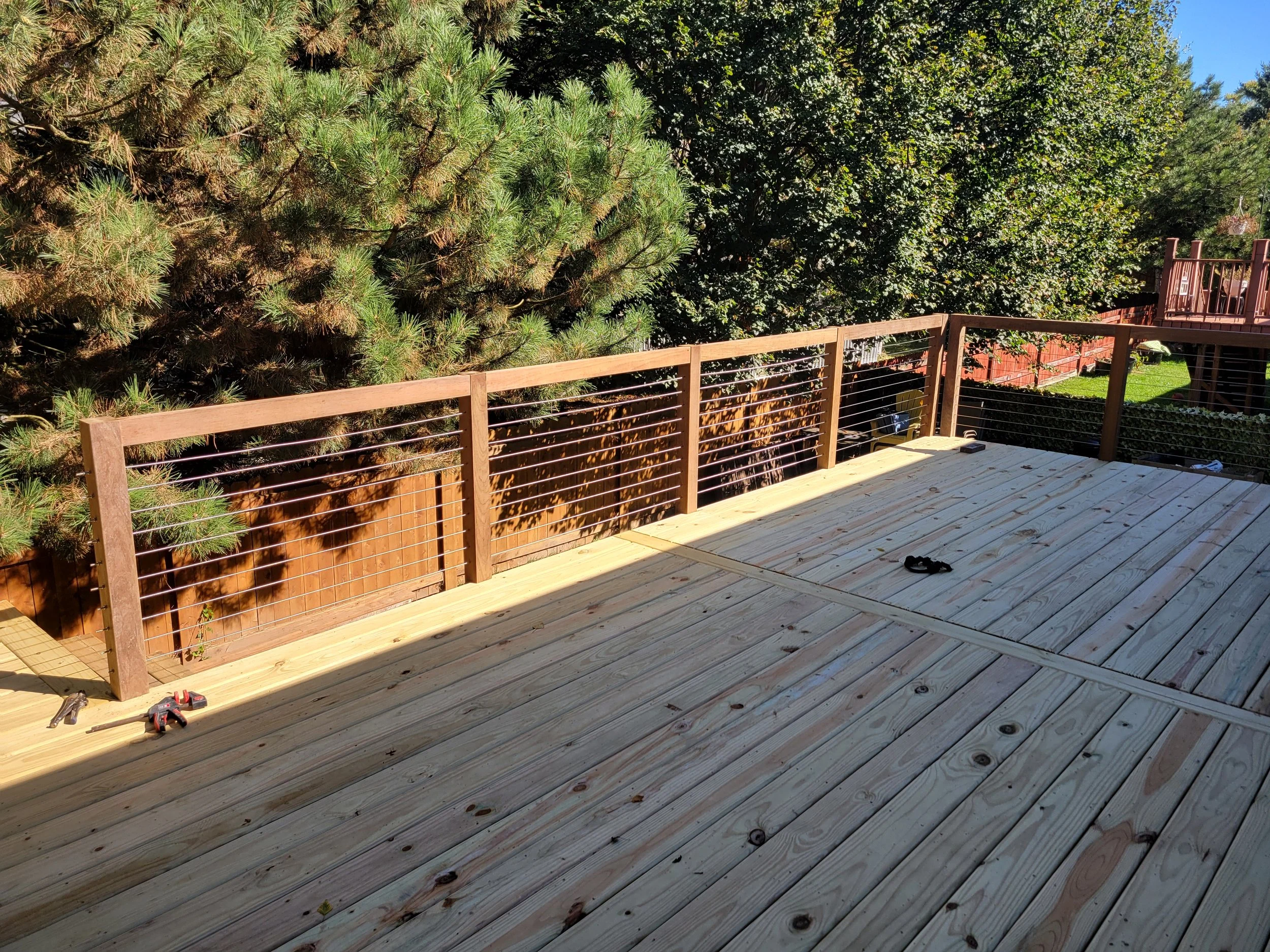 Newly built wooden deck with railing, outdoor tools on the floor, and a fence with trees in the background.