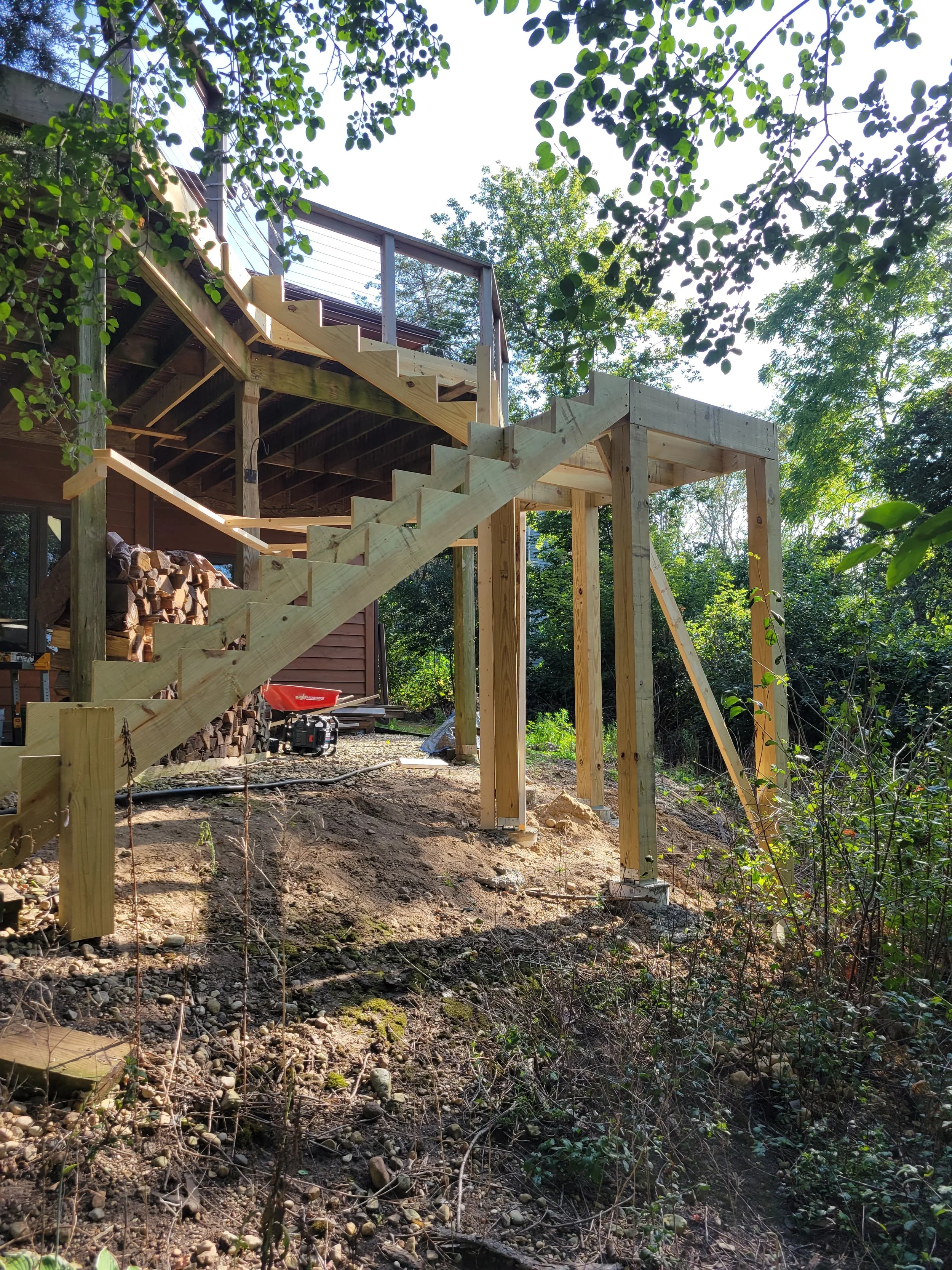 Wooden stairs under construction leading to a mid-level deck attached to a house, with trees and greenery in the background.
