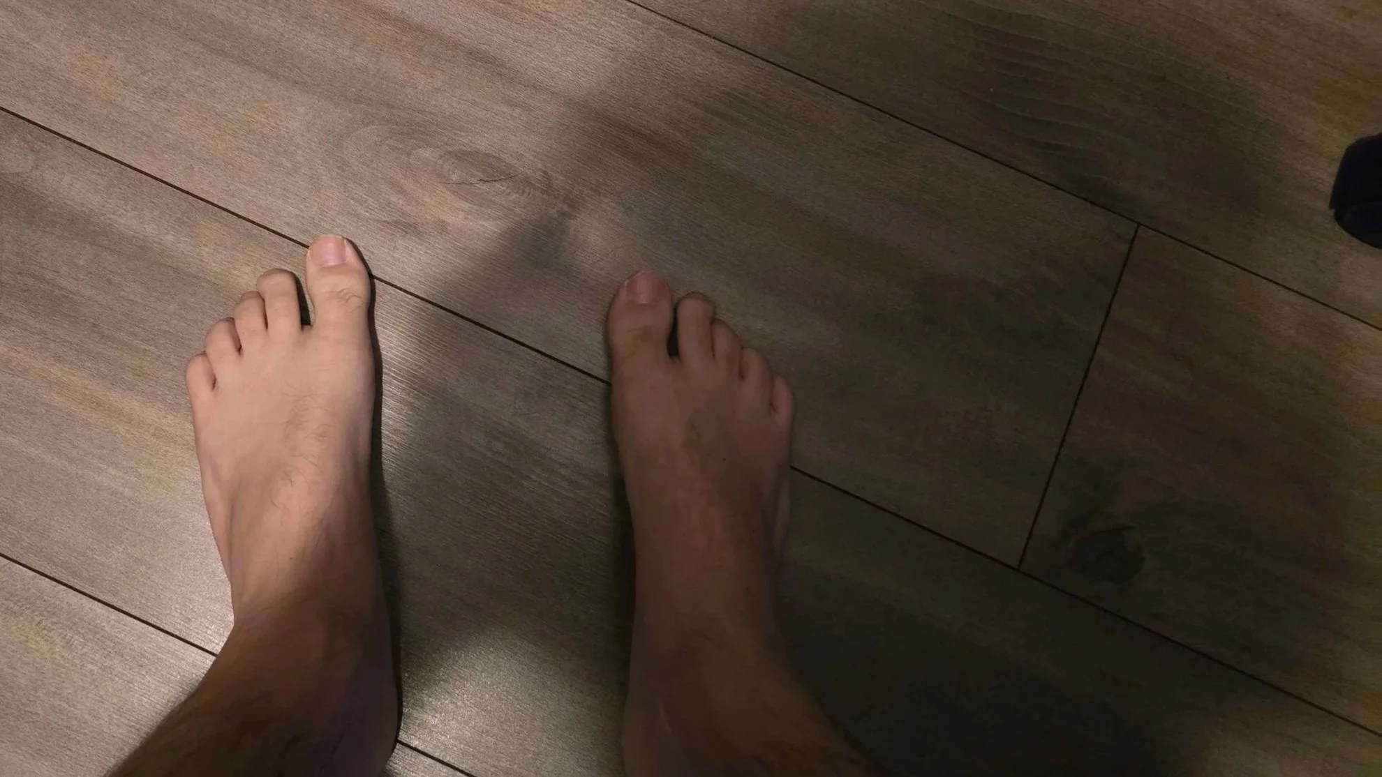 A person's bare feet standing on a wooden floor, with a dark sock visible on the right side of the image.