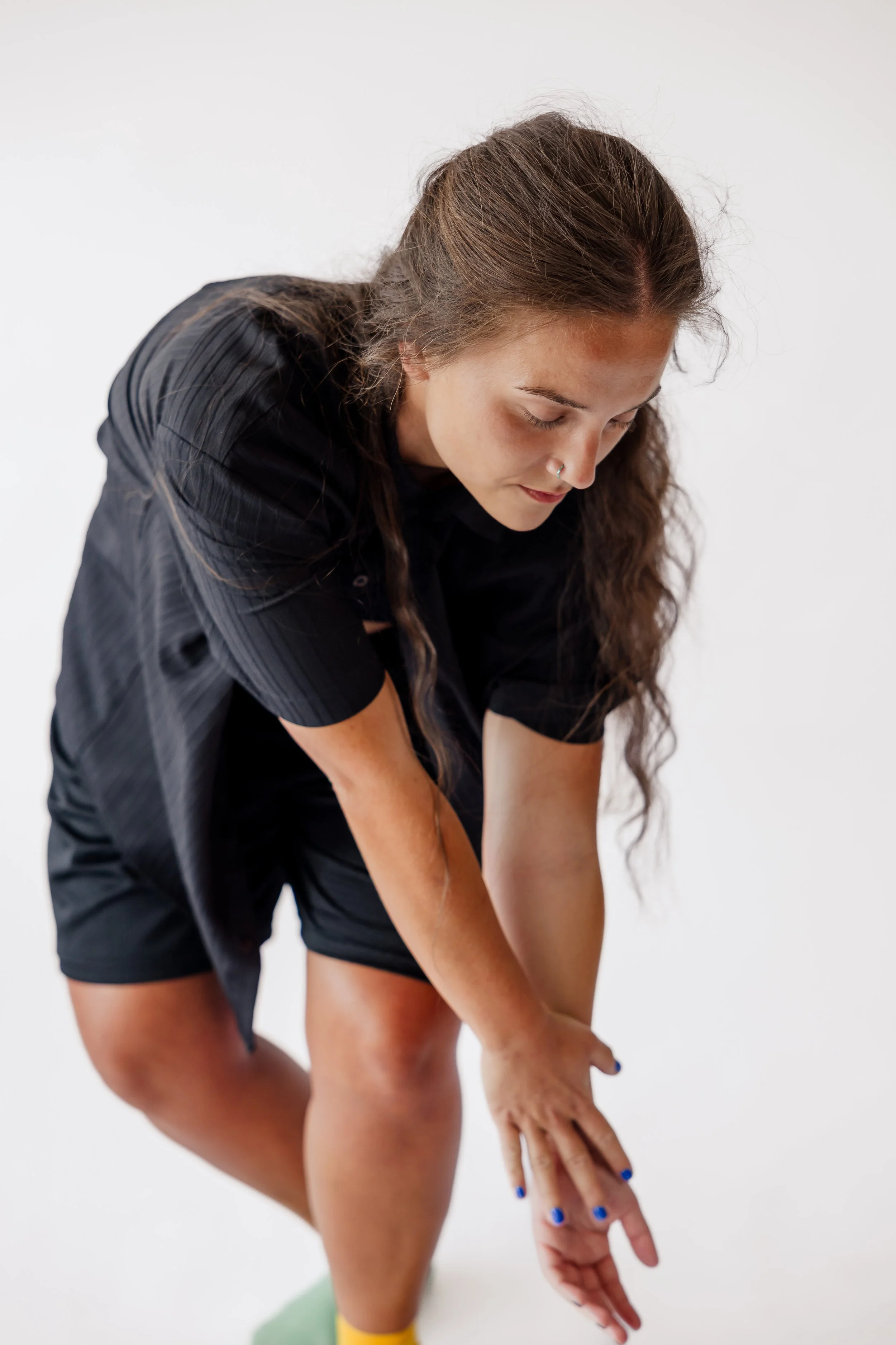 A young woman with long brown hair doing a stretching exercise in a black shirt and shorts, touching her toes with her right hand, against a plain white background.