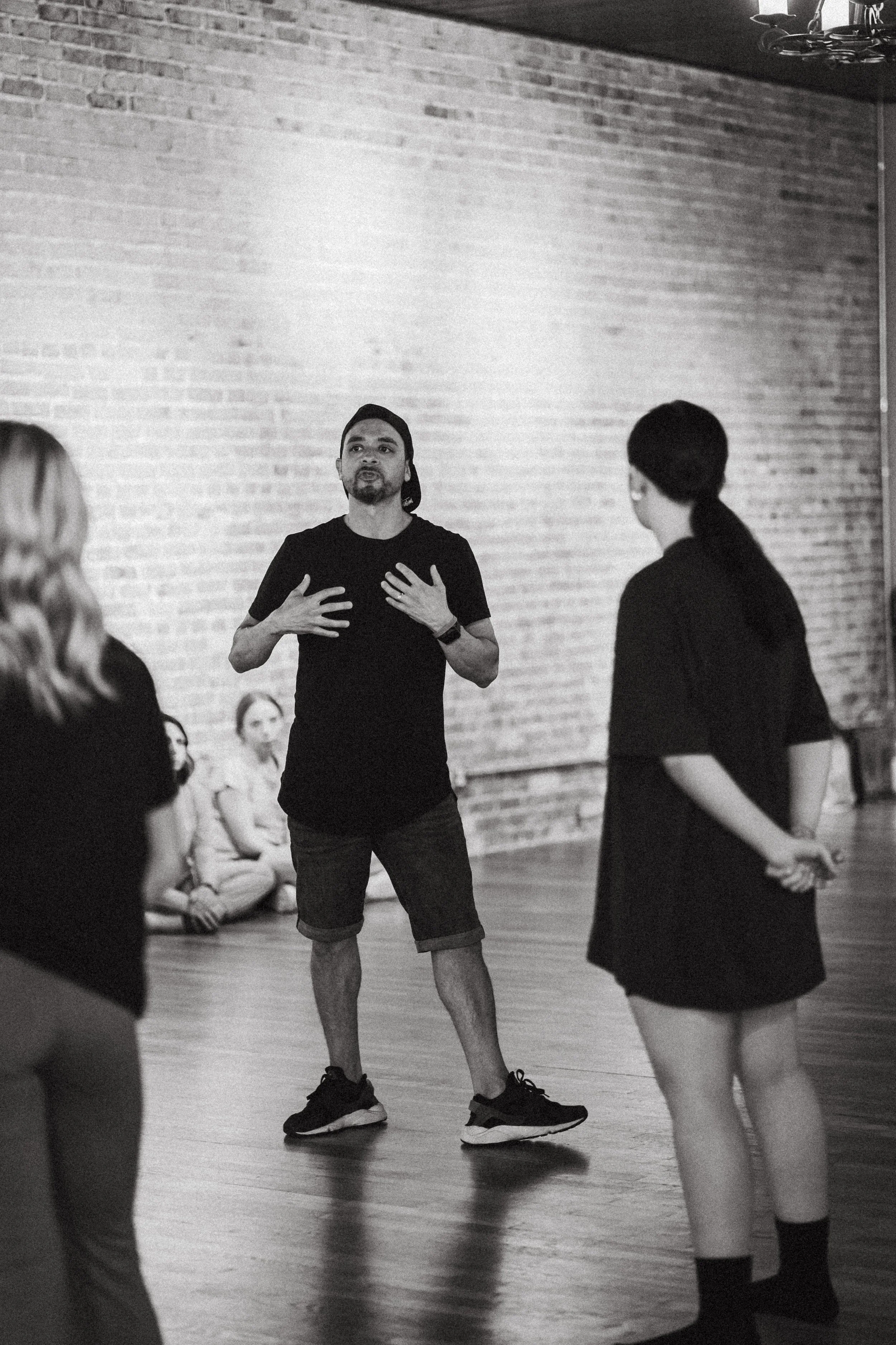A man with long hair, beard, and a black t-shirt speaking to a group of women in a dance studio with a brick wall background.