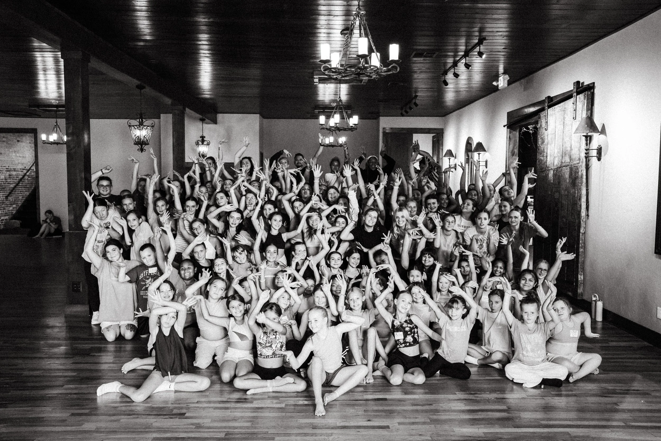 Group photo of children and adults indoors, posing with hands raised, in a room with wooden flooring, chandeliers, and rustic lighting.