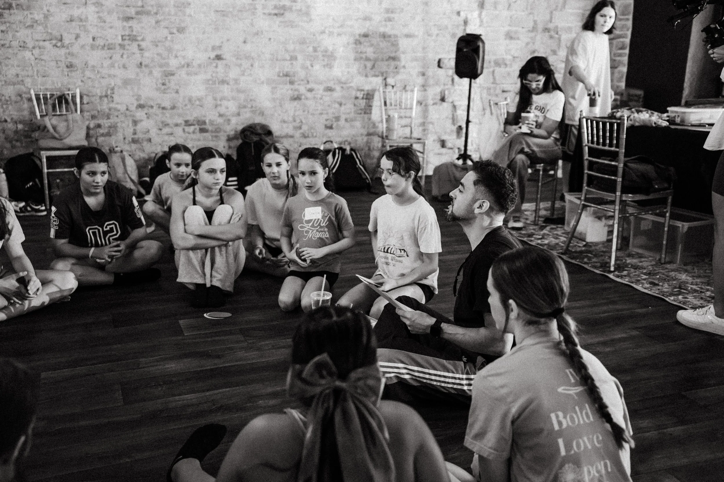 Group of young girls sitting on the floor in a circle, facing a young man who is reading from a book or paper, inside a room with exposed brick walls. Several chairs and backpacks are visible in the background.