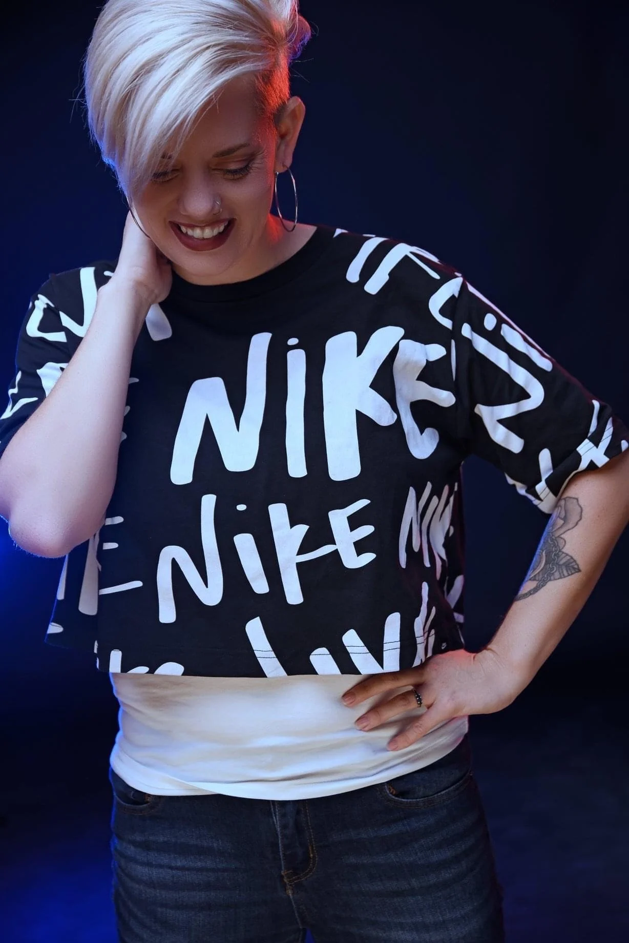 Young woman with short platinum blonde hair, wearing a black Nike t-shirt with white lettering, standing against a dark background with blue and red lighting, smiling and touching her neck.