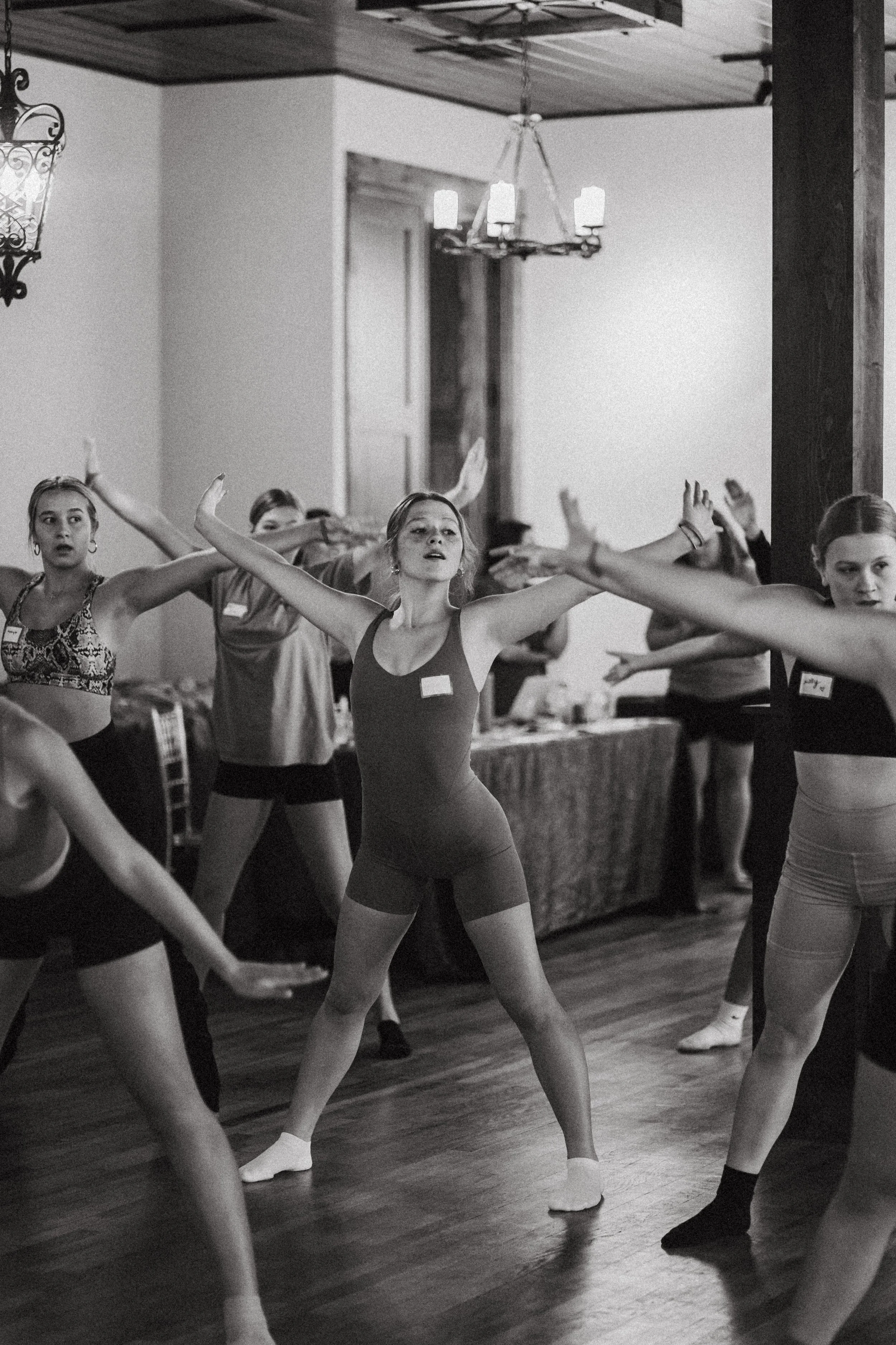 Ballet students practicing in a dance studio with wooden floors and a chandelier, black and white photograph.