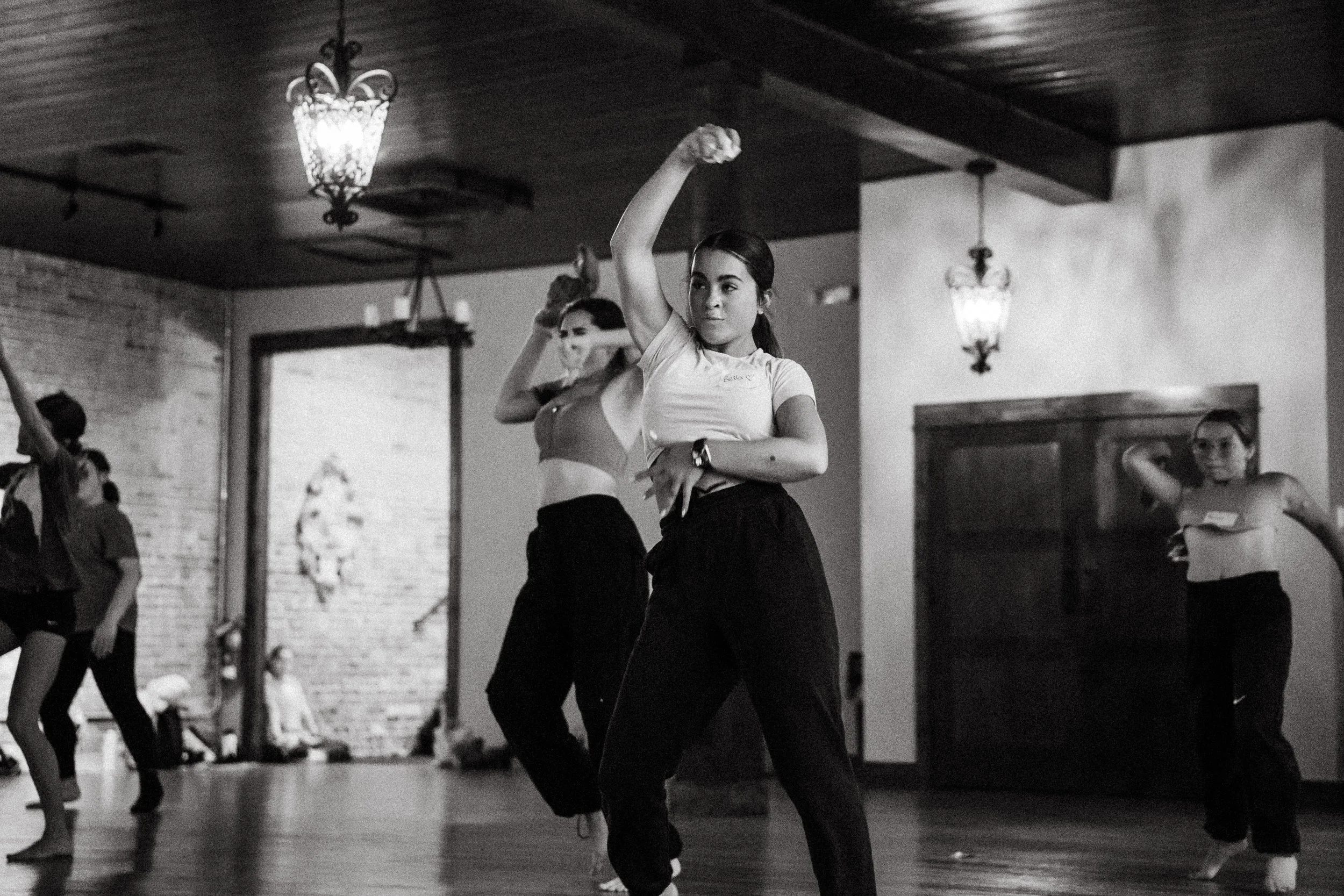 Young women practicing dance or fitness in a studio with brick walls and hanging lantern lights.