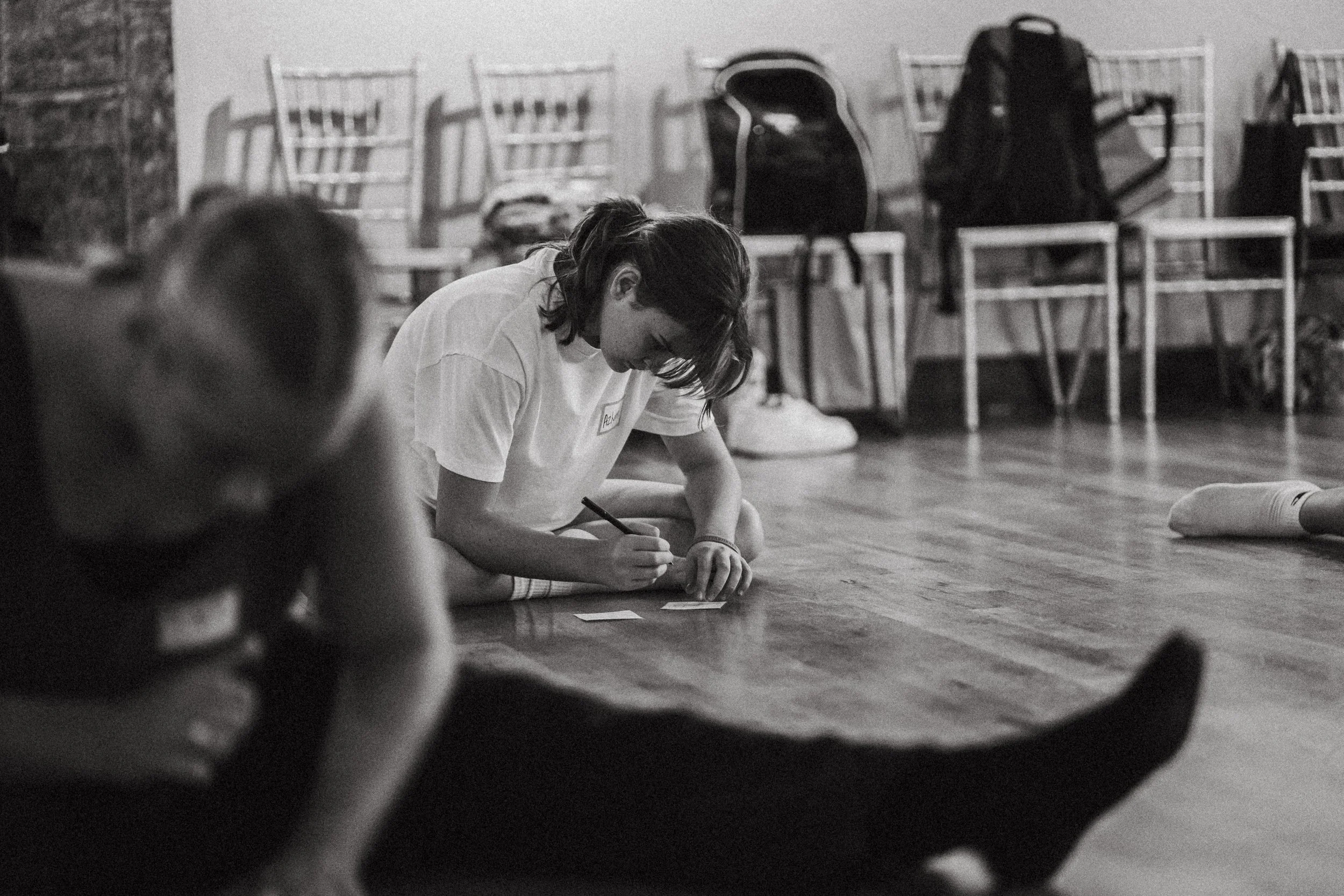 A person lying on the floor writing or drawing on small cards, with chairs and backpacks in the background.