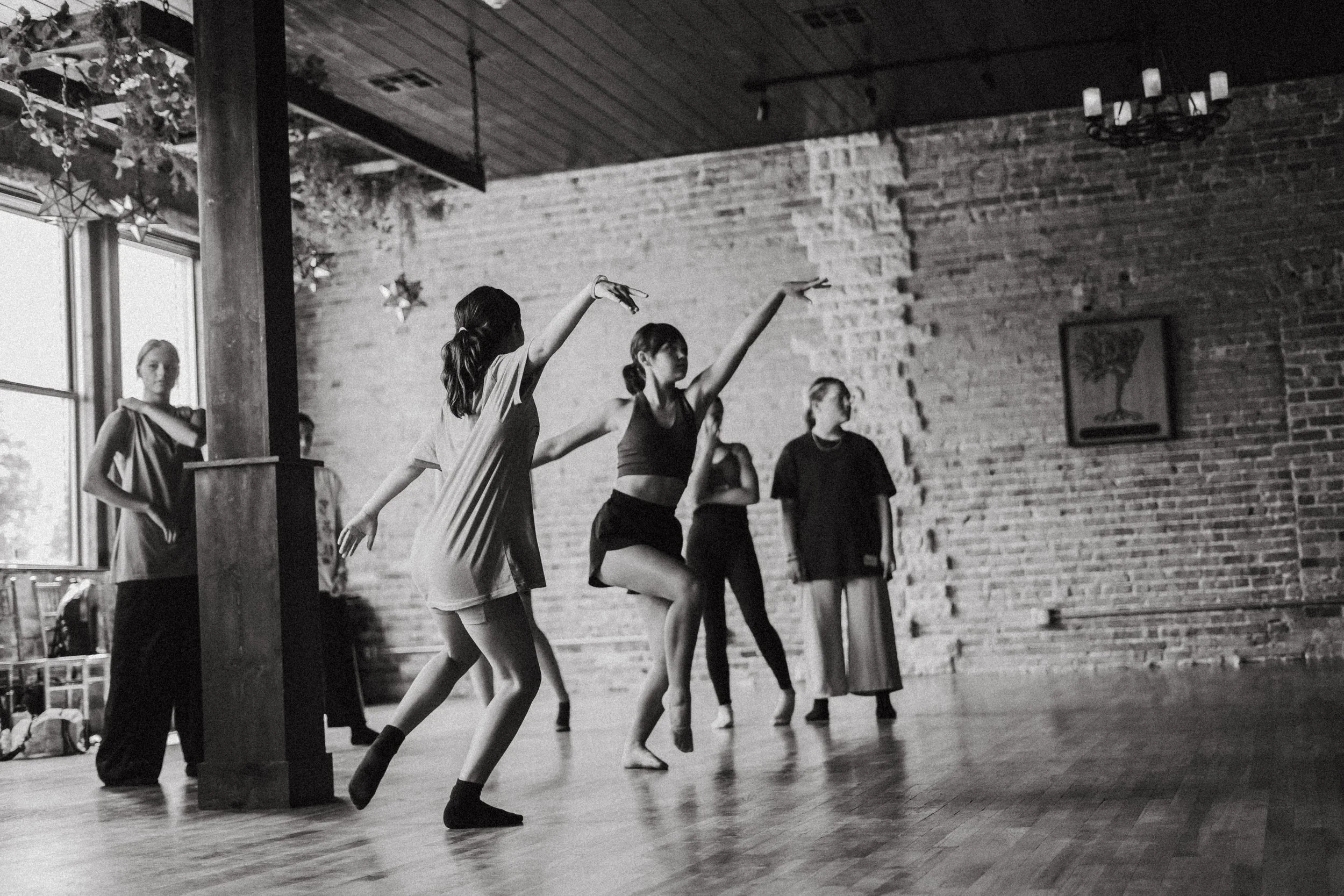 A group of young women practicing ballet in a studio with brick walls, large windows, and wooden floors, some performing dance moves.