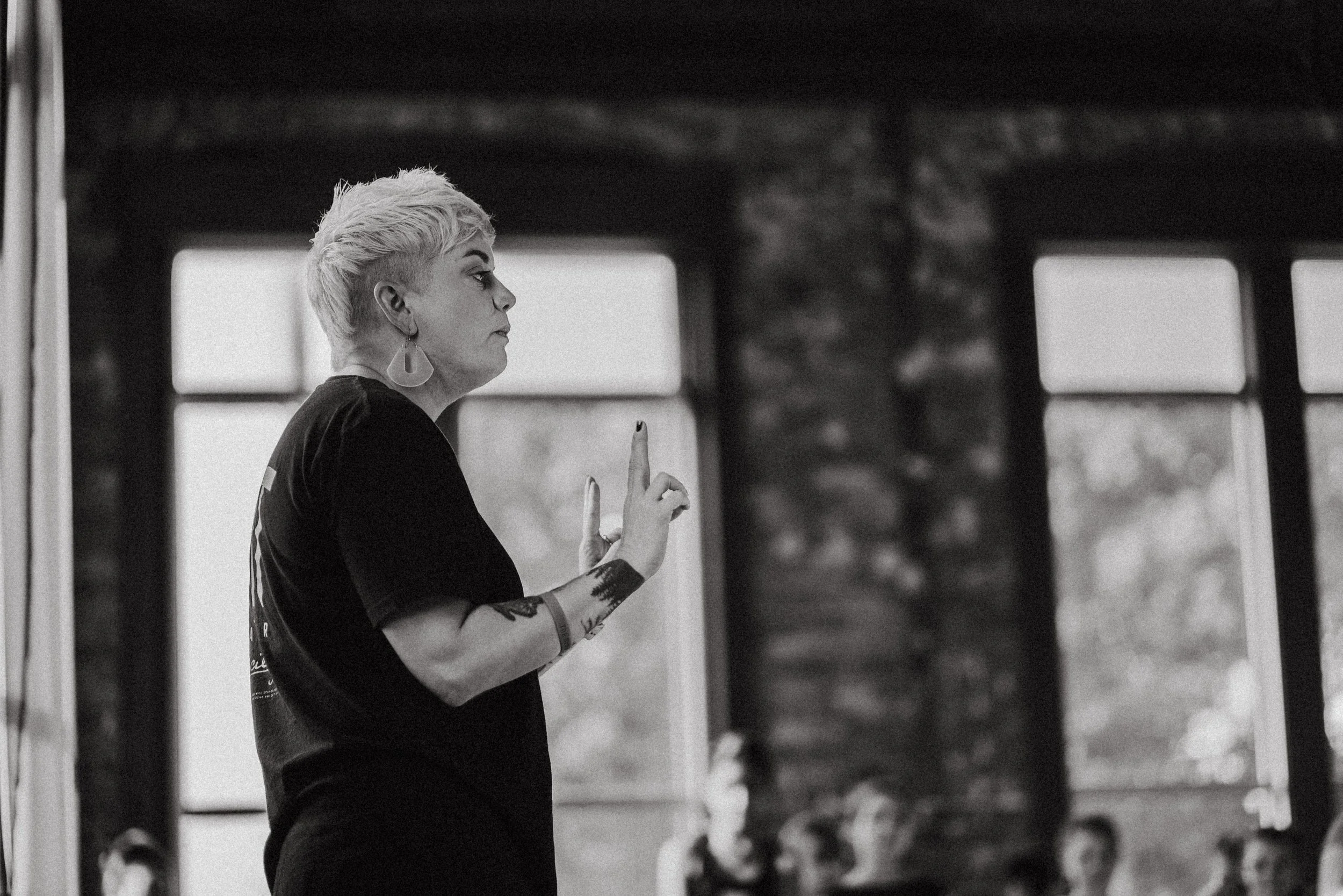 A woman with short, blonde hair, wearing earrings and a black shirt, stands inside a room with large windows. She is holding up her right hand with her index finger extended, possibly speaking to an audience.