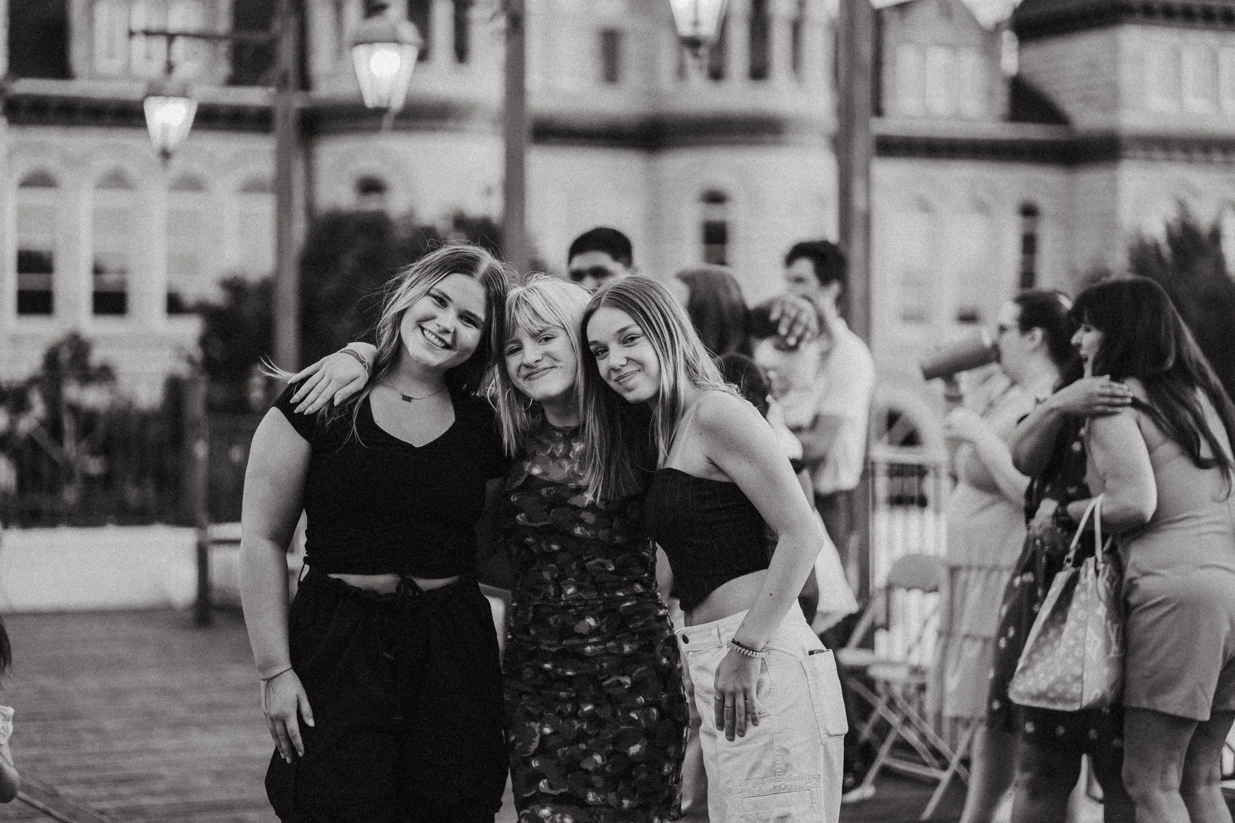 Three young women pose together smiling at an outdoor event, with adults and a large building in the background.