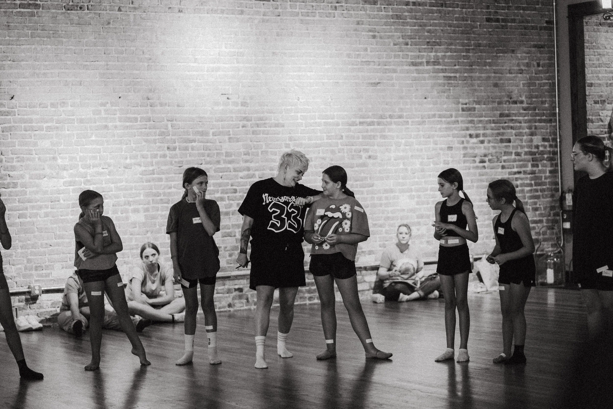 Group of young girls and a woman in a dance studio, with some sitting against a brick wall, participating in a dance class or rehearsal.