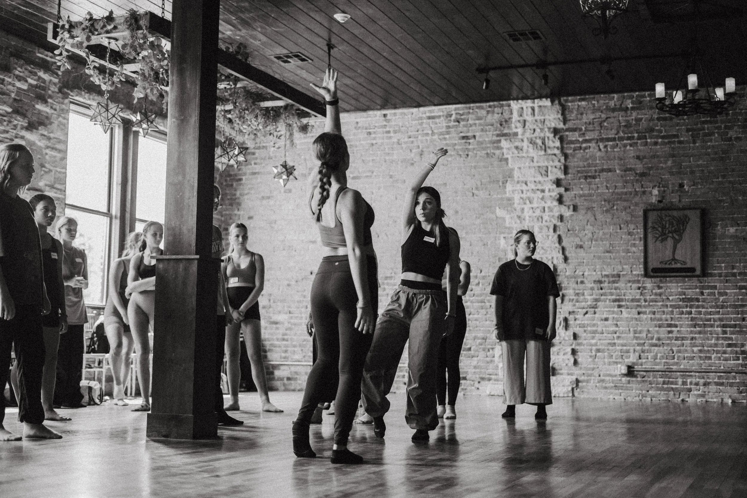 Women practicing dance in a studio with brick walls and wooden flooring, one woman demonstrating moves while others watch.