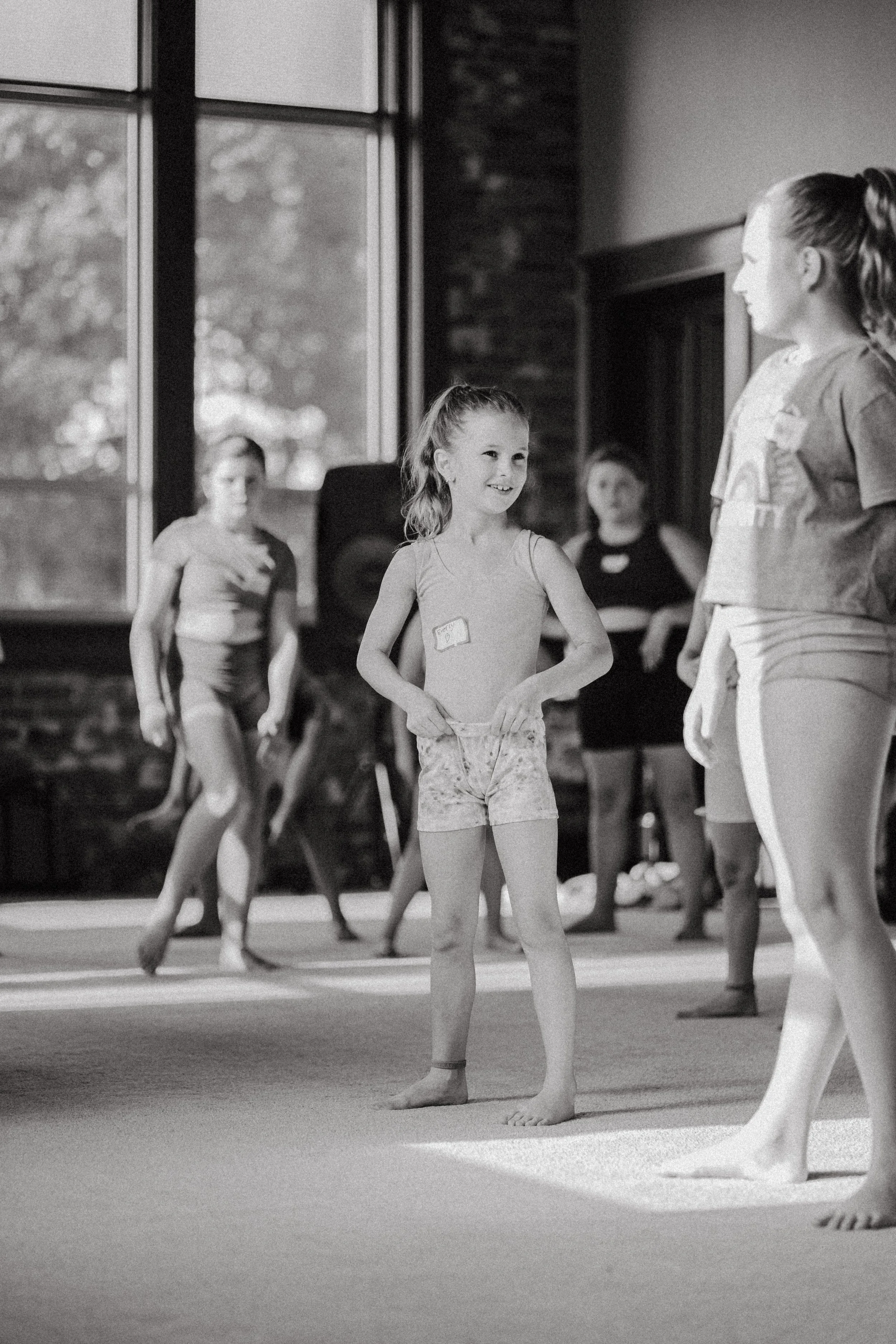 Children participating in a dance or gymnastics class indoors, with one girl smiling at another, other children and an instructor visible in the background.