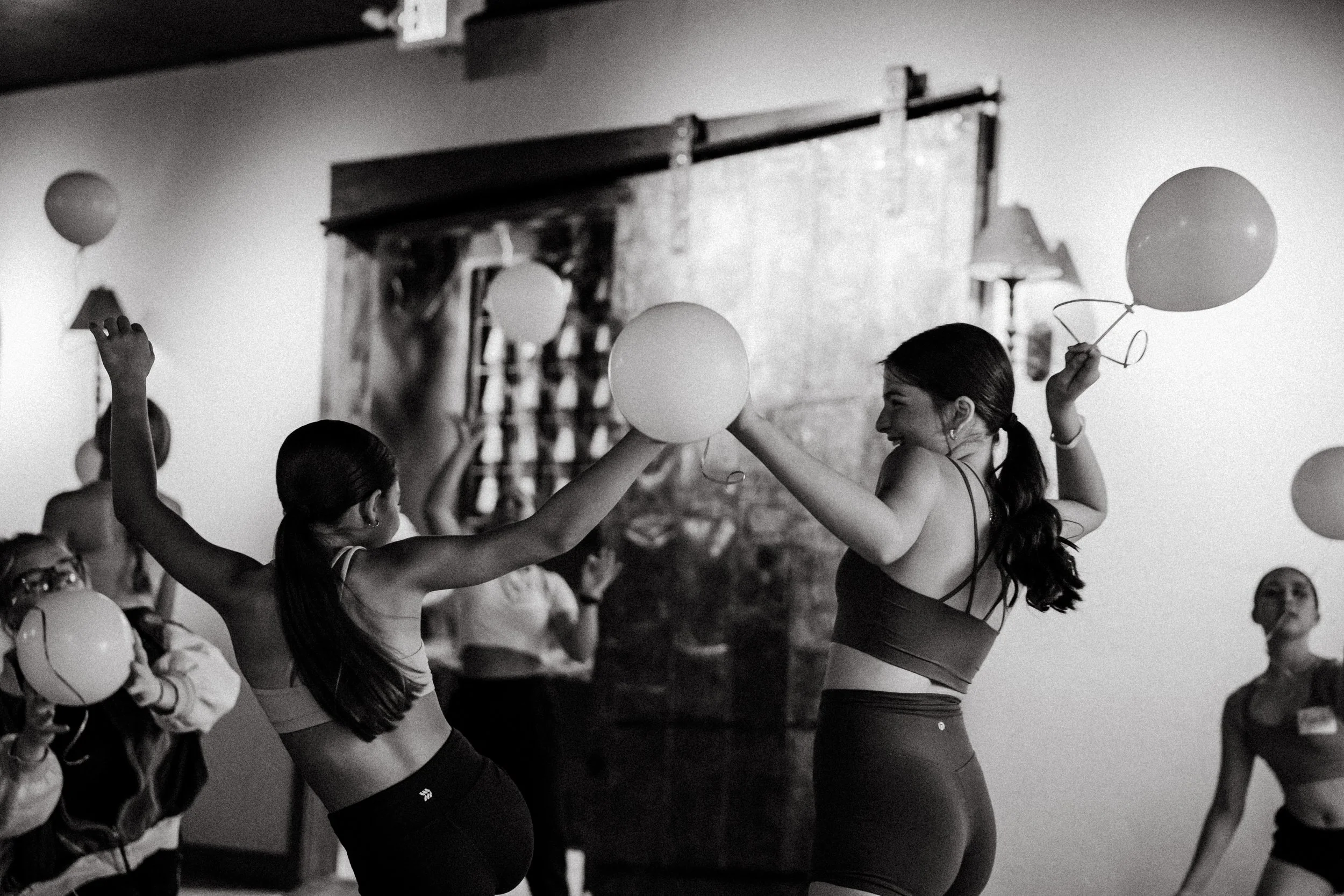 Two women dance indoors, holding balloons and smiling, with other women in the background. Black and white photo.