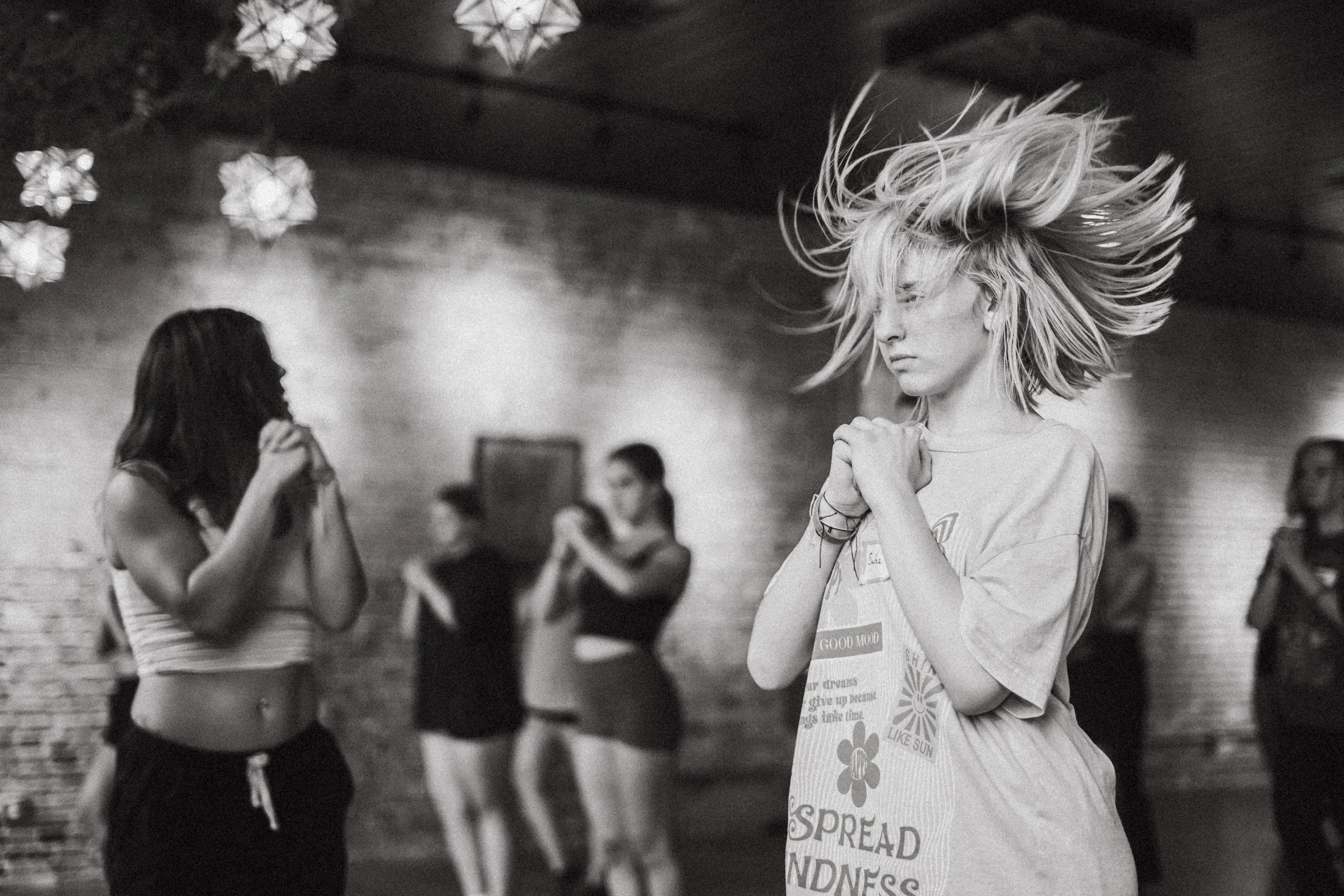 A group of young women dancing and practicing ballet or dance moves in a studio with brick walls and hanging paper decorations.