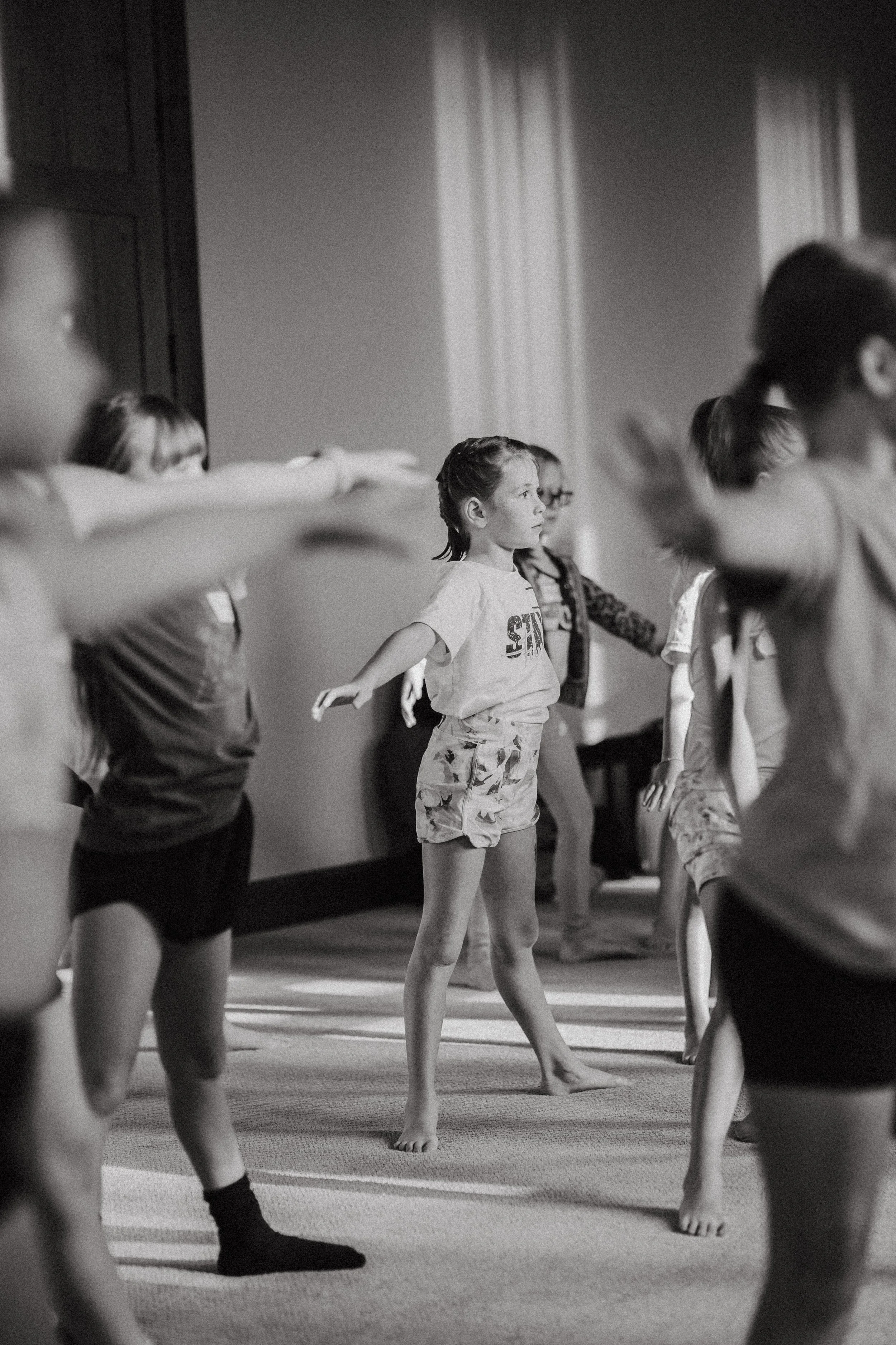 A group of children participating in a dance or exercise class, standing on one leg with arms extended outward, indoors with curtains and wooden paneling visible in the background.