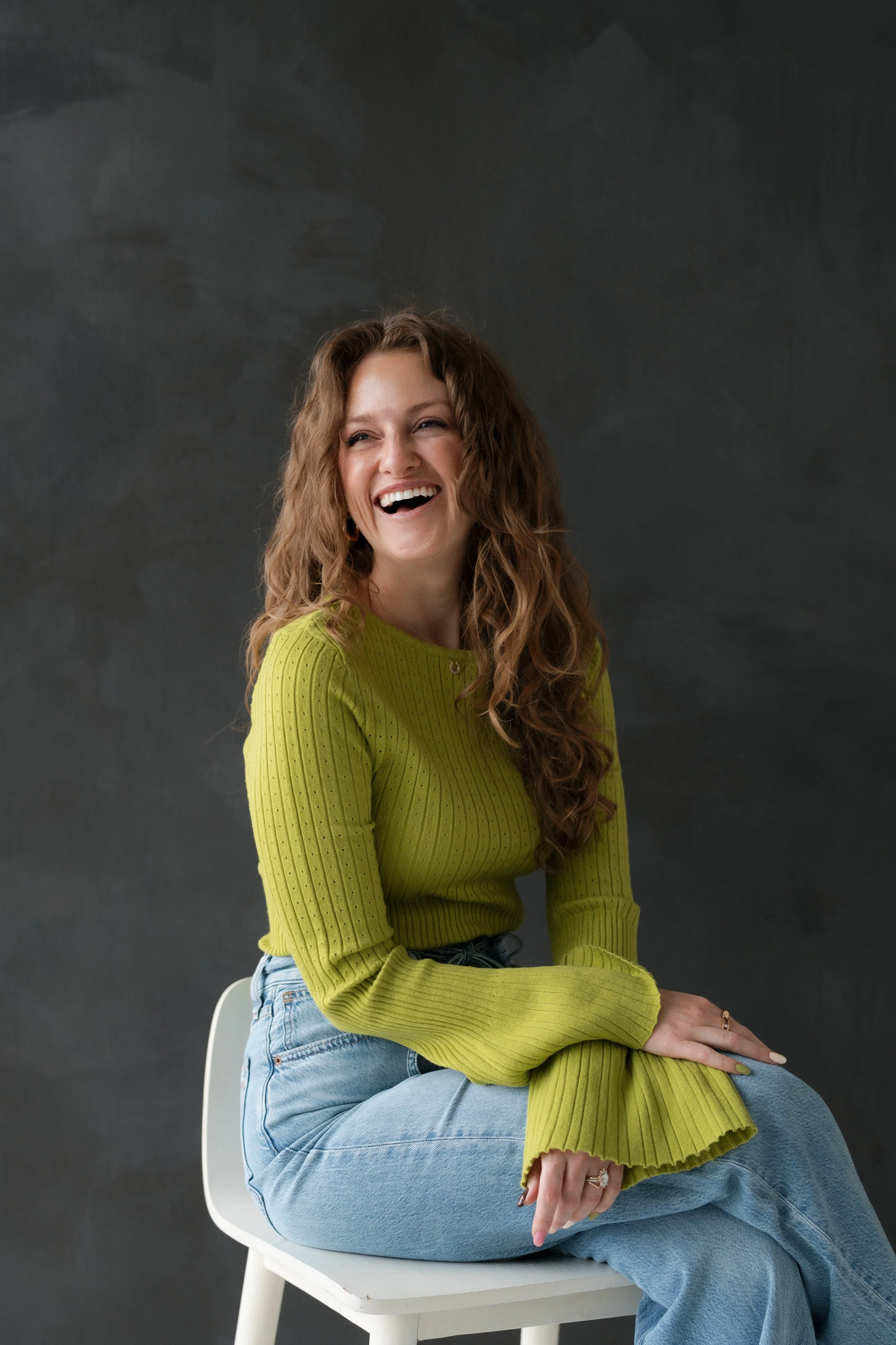 A smiling woman with long curly hair, wearing a neon green ribbed sweater and light blue jeans, sitting on a white chair against a dark background.