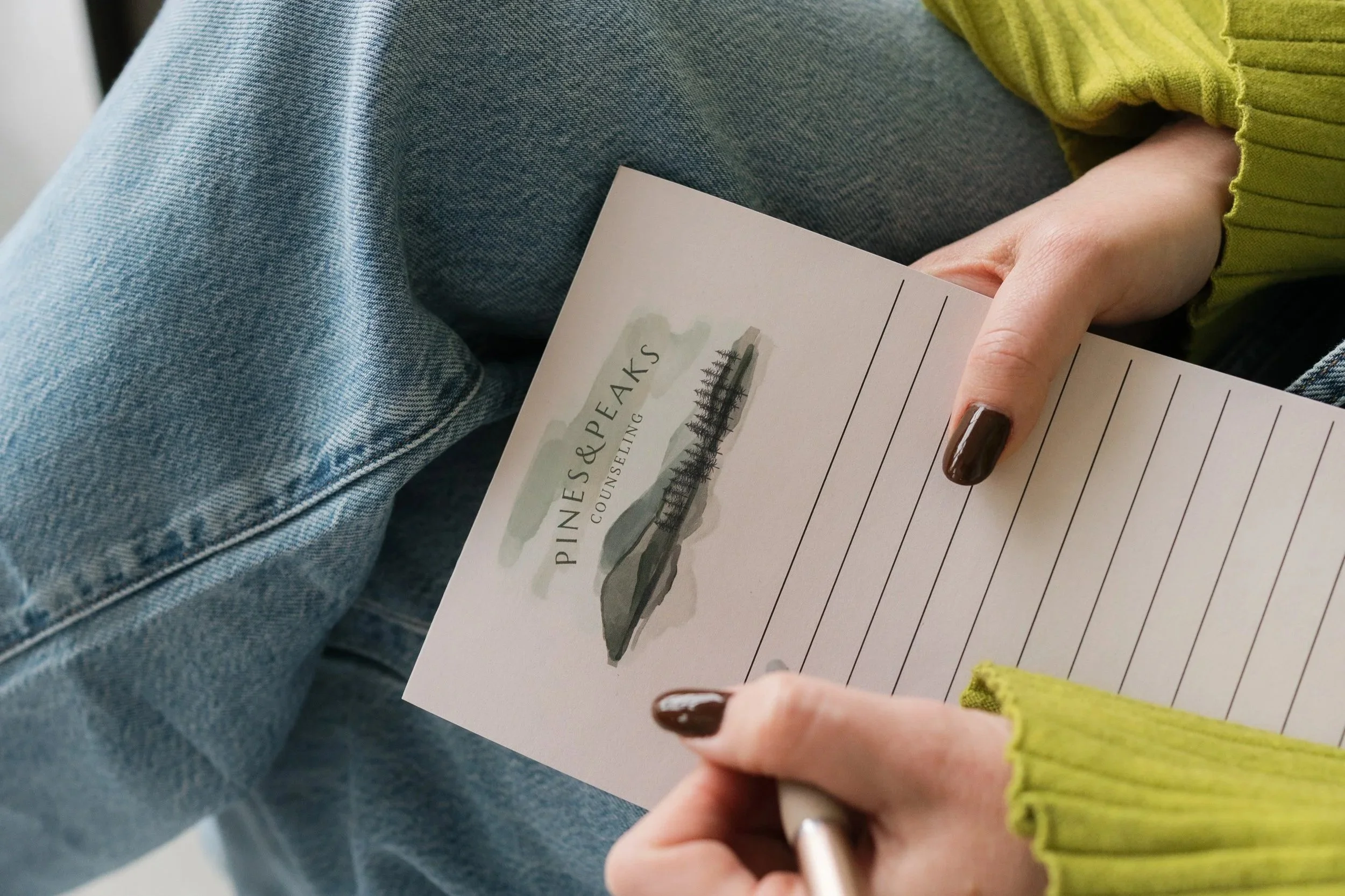 Person holding a pen and a printed piece of paper with a watercolor illustration of a pier, with blue jeans and a green long-sleeve shirt.