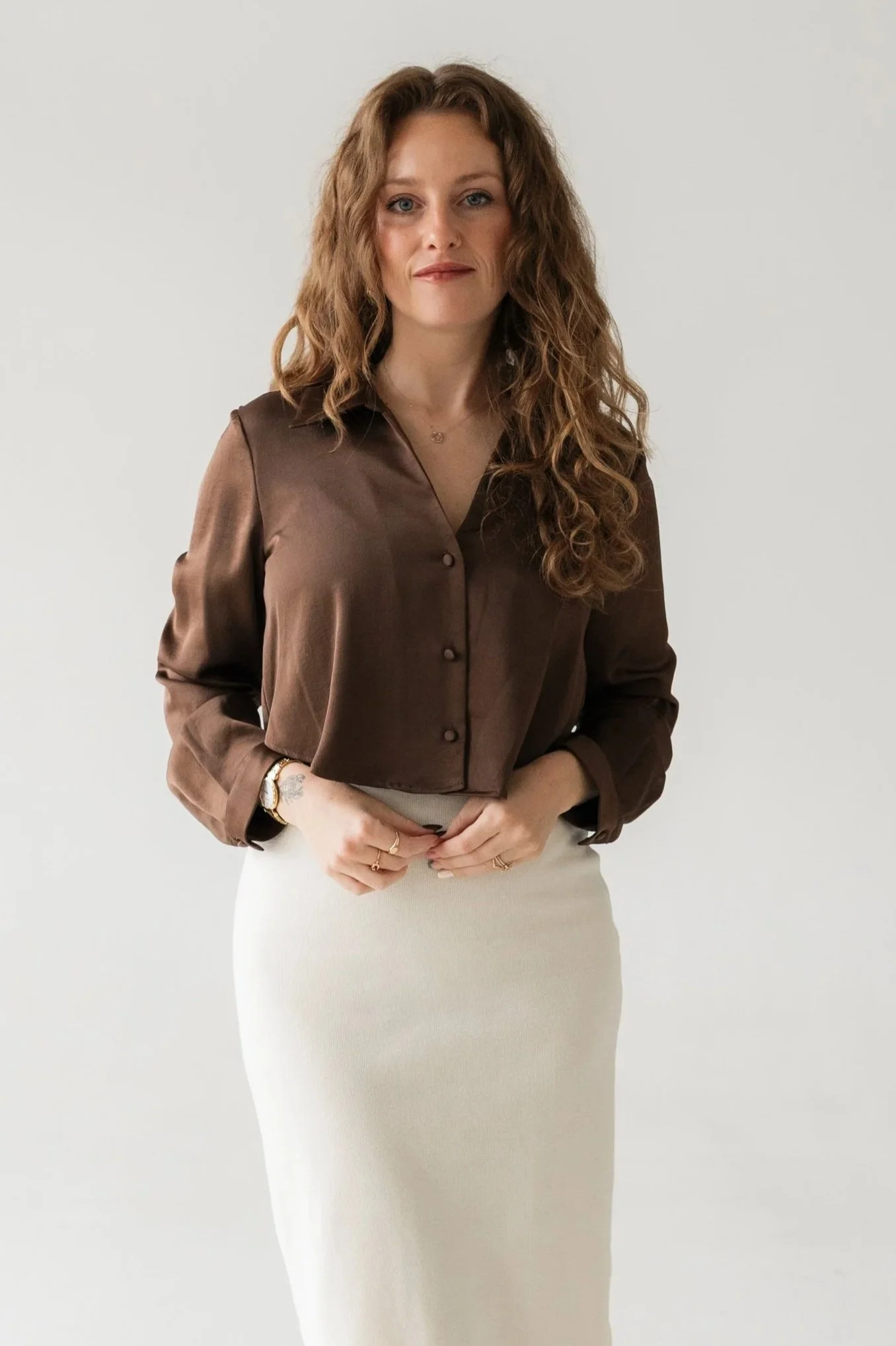 A woman with long wavy hair wearing a brown blazer and a white skirt, standing against a plain light background.