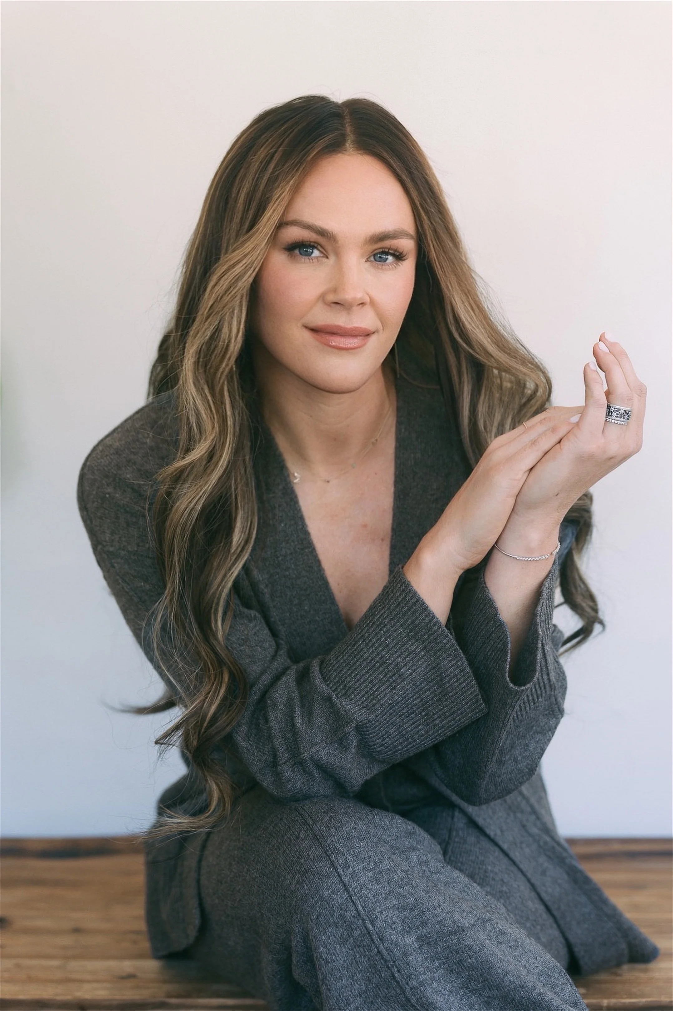 Svetlana Ishchenko of Vera Nova Therapy with long wavy hair, wearing a gray suit, sitting on a wooden floor against a plain white background, looking at the camera with a slight smile.