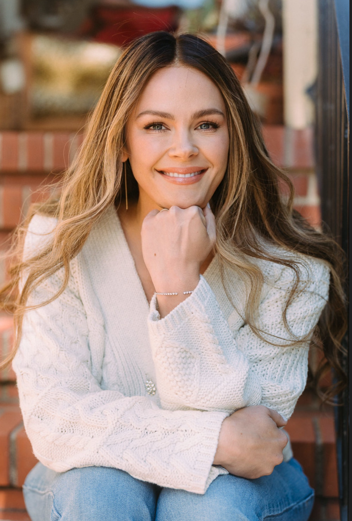 Svetlana Ishchenko of Vera Nova Therapy Ishchenko with long, wavy, light brown hair and blue eyes sitting outdoors on brick stairs, smiling and resting her chin on her hand, wearing a white knitted sweater and blue jeans.
