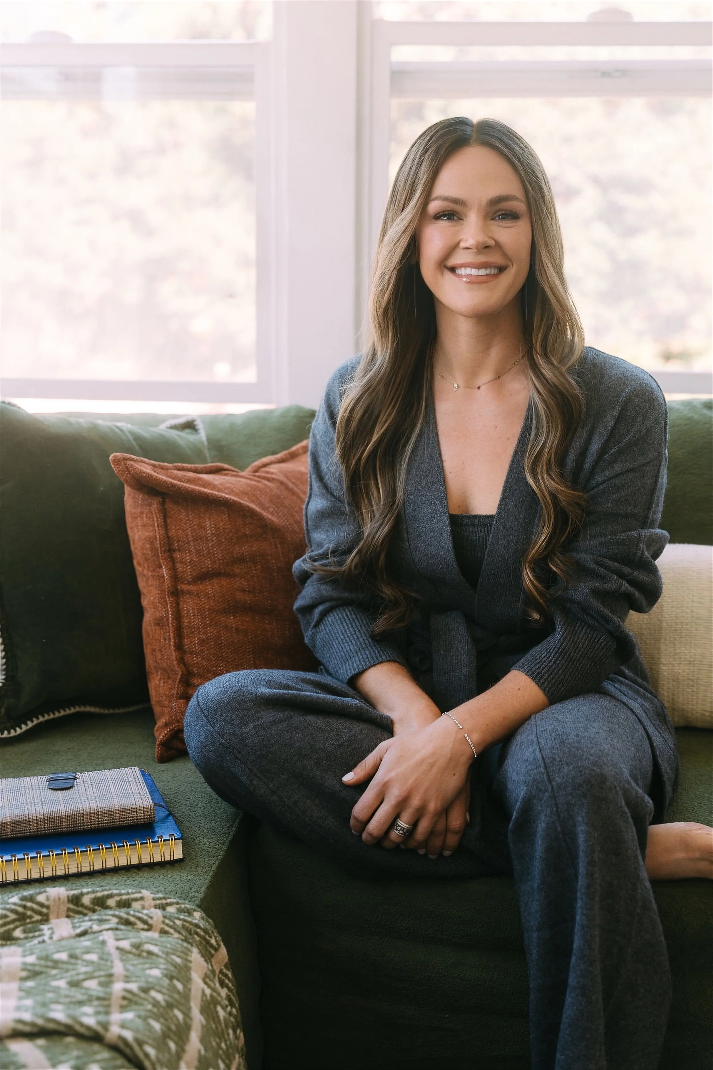 Svetlana Ishchenko of Vera Nova Therapy with long wavy hair, smiling, sitting cross-legged on a green couch in front of a window, wearing a dark gray sweater and pants, with notebooks and pillows nearby.