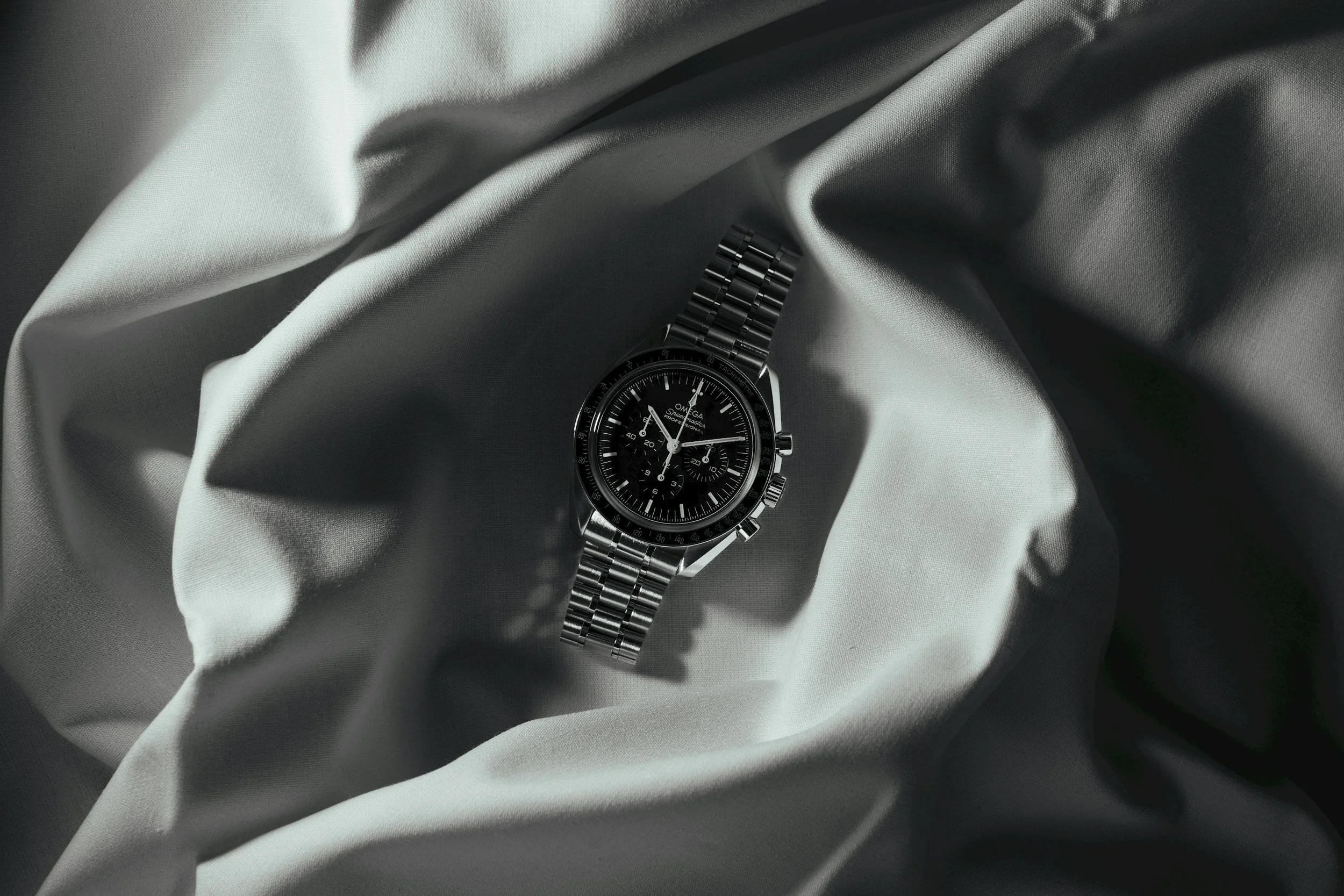 A black and silver Omega wristwatch with a metal band, lying on white fabric with folds and shadows.