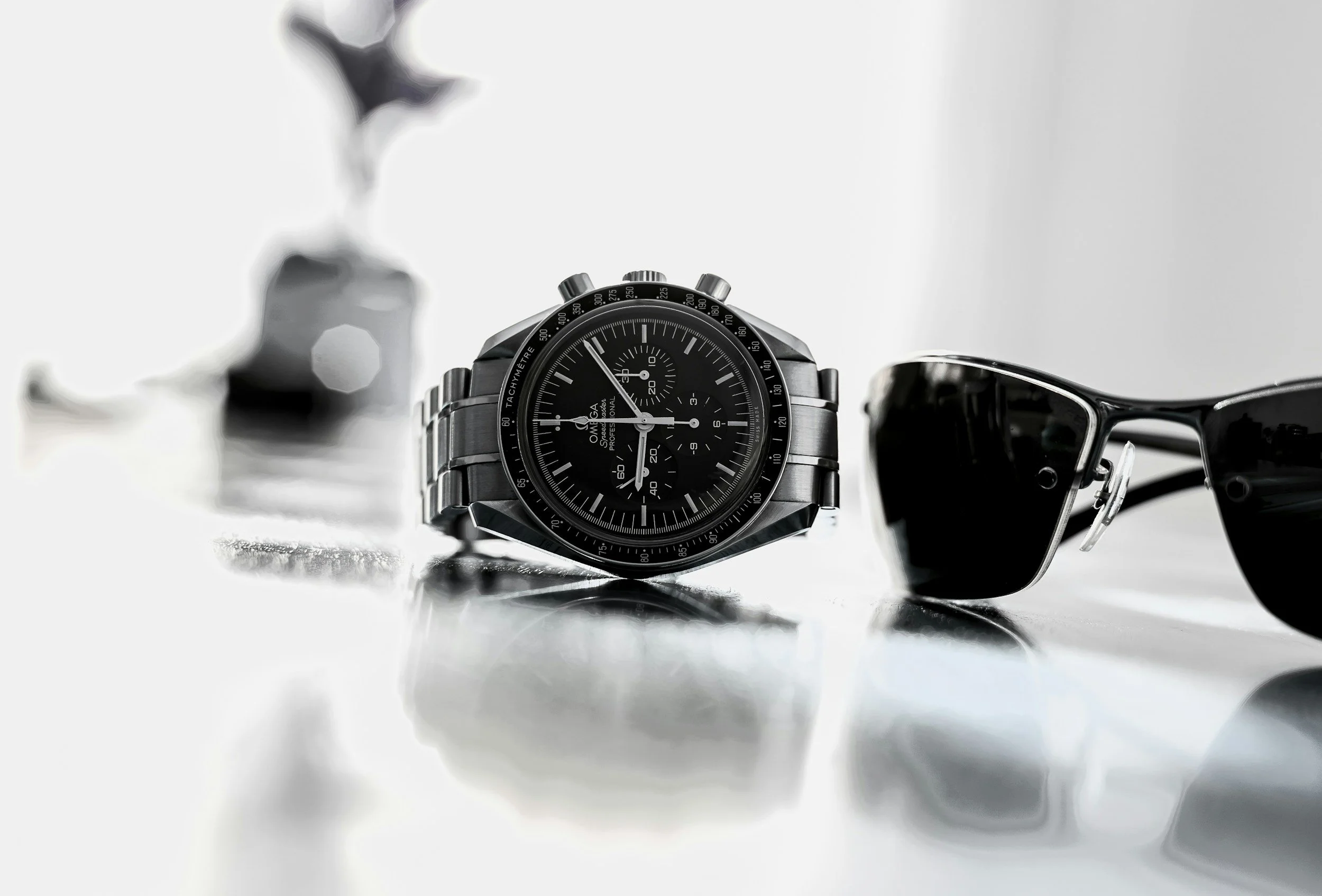 A black Omega Speedmaster wristwatch with a metal bracelet, placed on a reflective surface. To the right, there are black sunglasses with a reflective surface. The background is plain and minimalist.