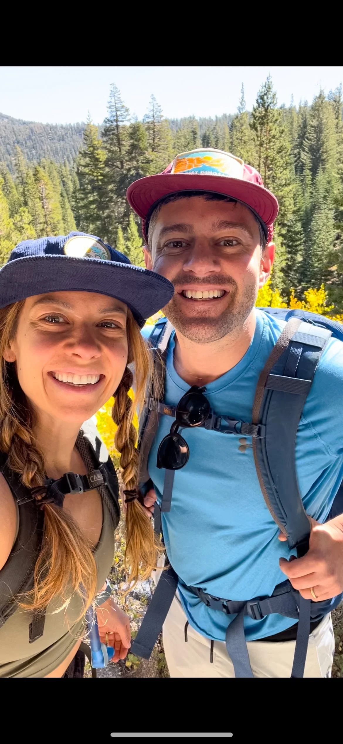 Two smiling hikers taking a selfie outdoors with a forest and mountains in the background.