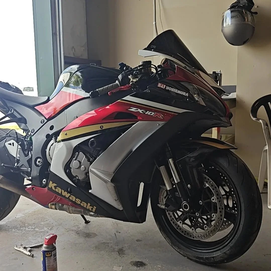 A Kawasaki ZX10R sport motorcycle parked indoors near a shop entrance with a black windshield and a red, black, and white color scheme. There is a helmet hanging on the wall and a red and white brake caliper on the front wheel.