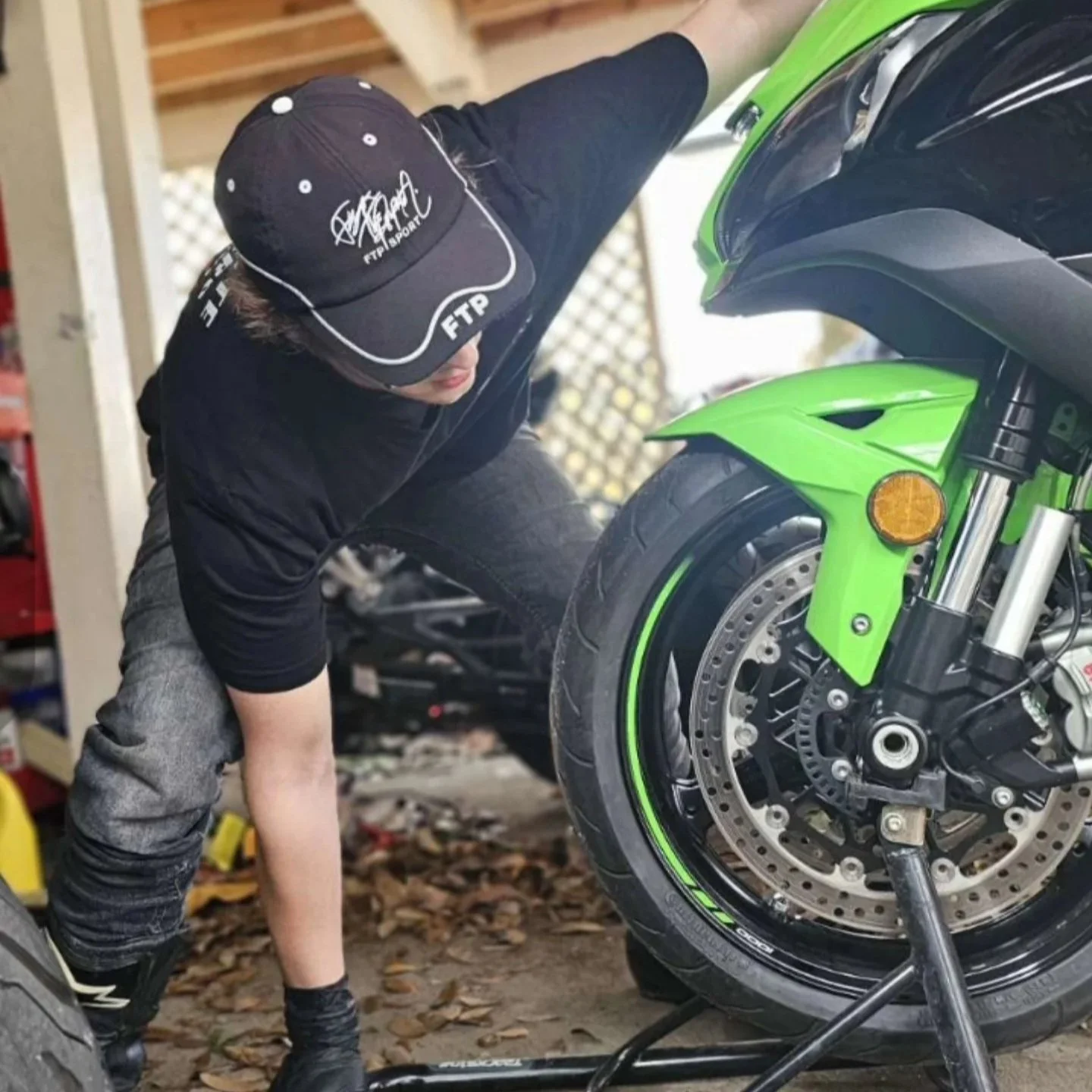 A person wearing a black cap, black shirt, and grey jeans working on a green motorcycle indoors.