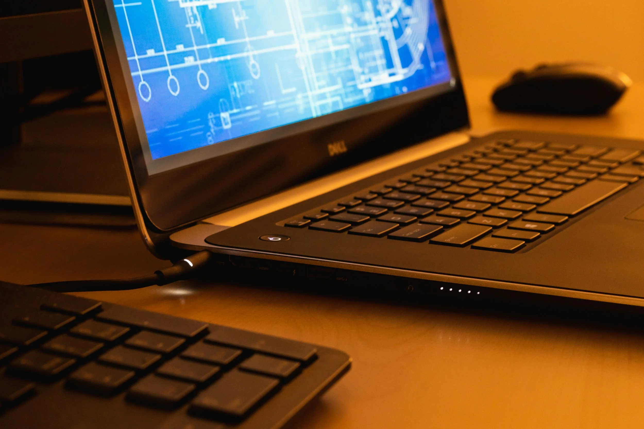 Close-up of a laptop computer displaying a blue technical or architectural blueprint on its screen, connected to a power source, with a wireless mouse in the background.
