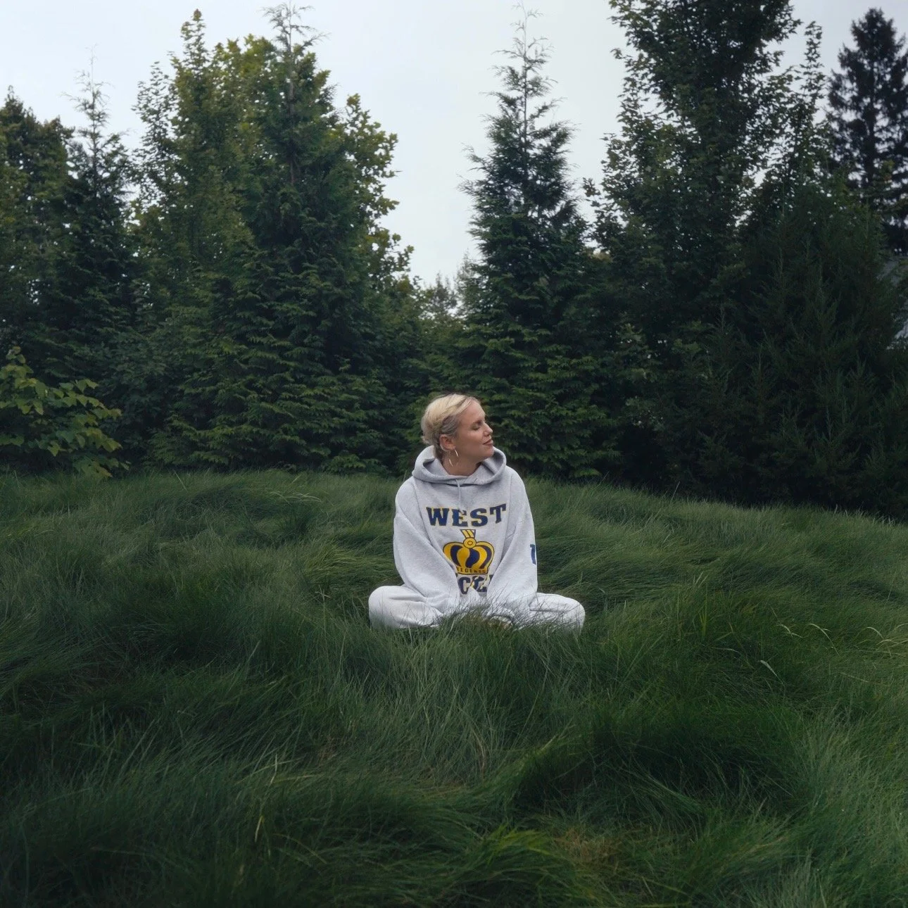 A woman sitting cross-legged on lush green grass in a forest clearing, wearing a gray hoodie with 'WEST' and a crown logo on it, surrounded by tall pine trees.