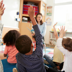 Children in a classroom raising their hands while a teacher stands at the front near a whiteboard.