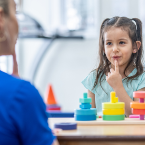 A young girl with pigtails touching her lips while playing with colorful stacking toys, sitting across from an adult.