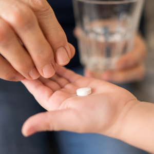 A person hand holding a pill with another hand receiving it, with a glass of water in the background.