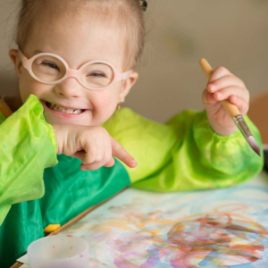A young child with glasses smiling and holding a paintbrush while painting at a table.