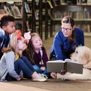 A woman reading a book to a group of children and a dog in a library.
