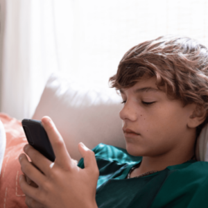 Teen boy with brown hair sitting on a bed, holding and looking at a smartphone.