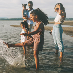 A family enjoying time at the beach, with a girl kicking water, a man holding a child, and two women standing nearby.