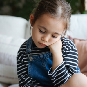 A young girl with her head resting on her hand, looking down pensively, wearing a striped shirt and denim overalls.