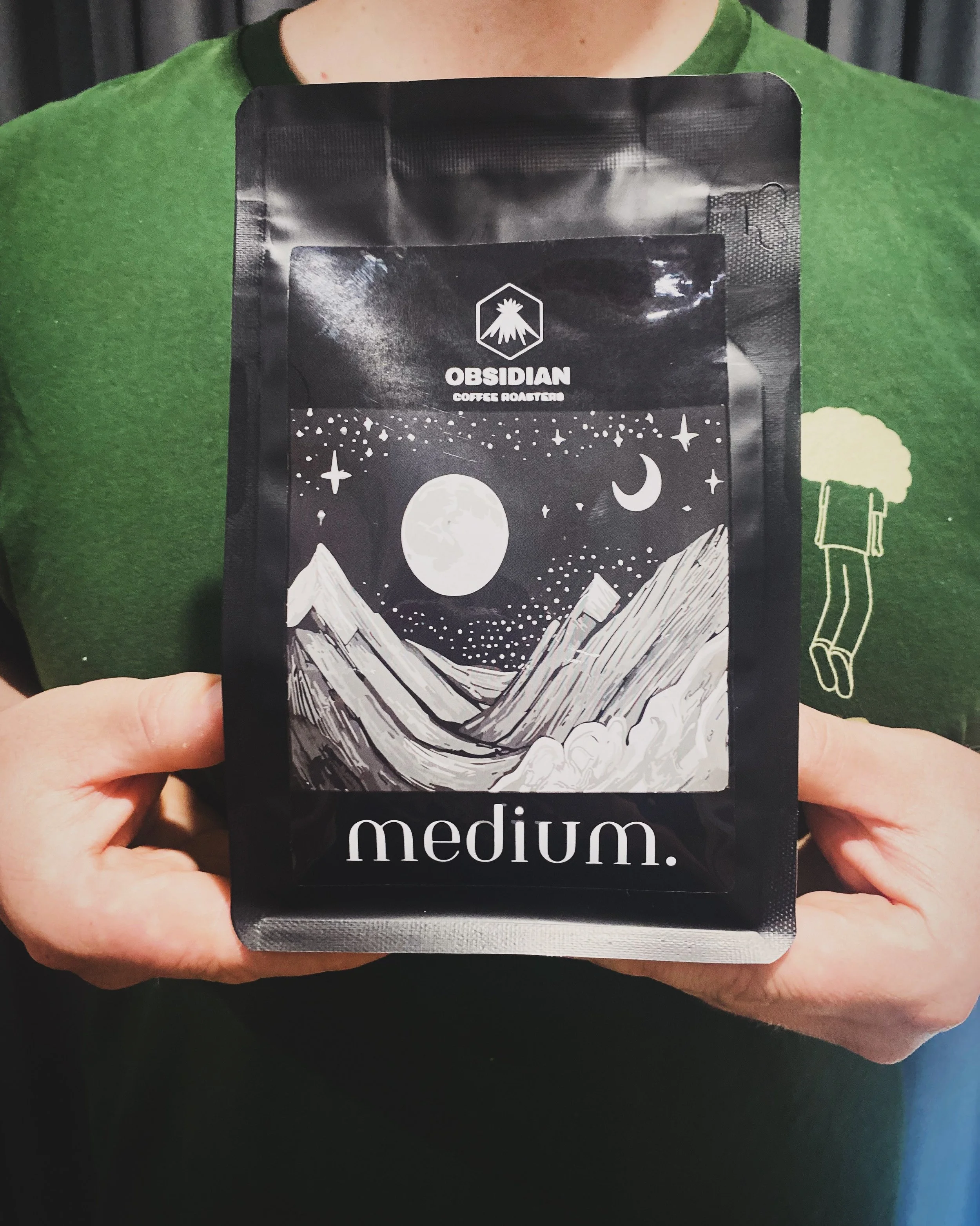 Person holding a black bag of Obsidian coffee roaster labeled 'medium' in front of a green shirt with a graphic of a beer mug.