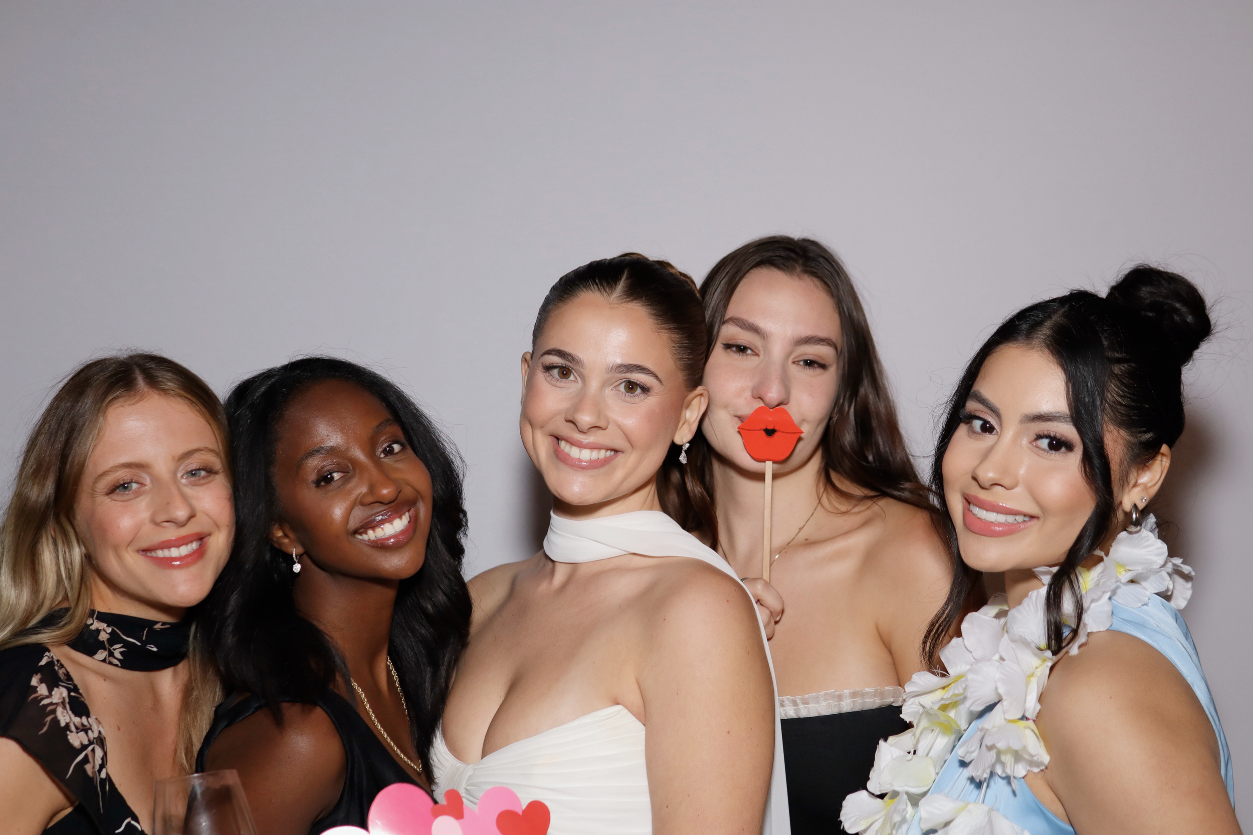 Group of five women smiling, celebrating, with one woman holding a paper lips prop, and another with a flower lei.