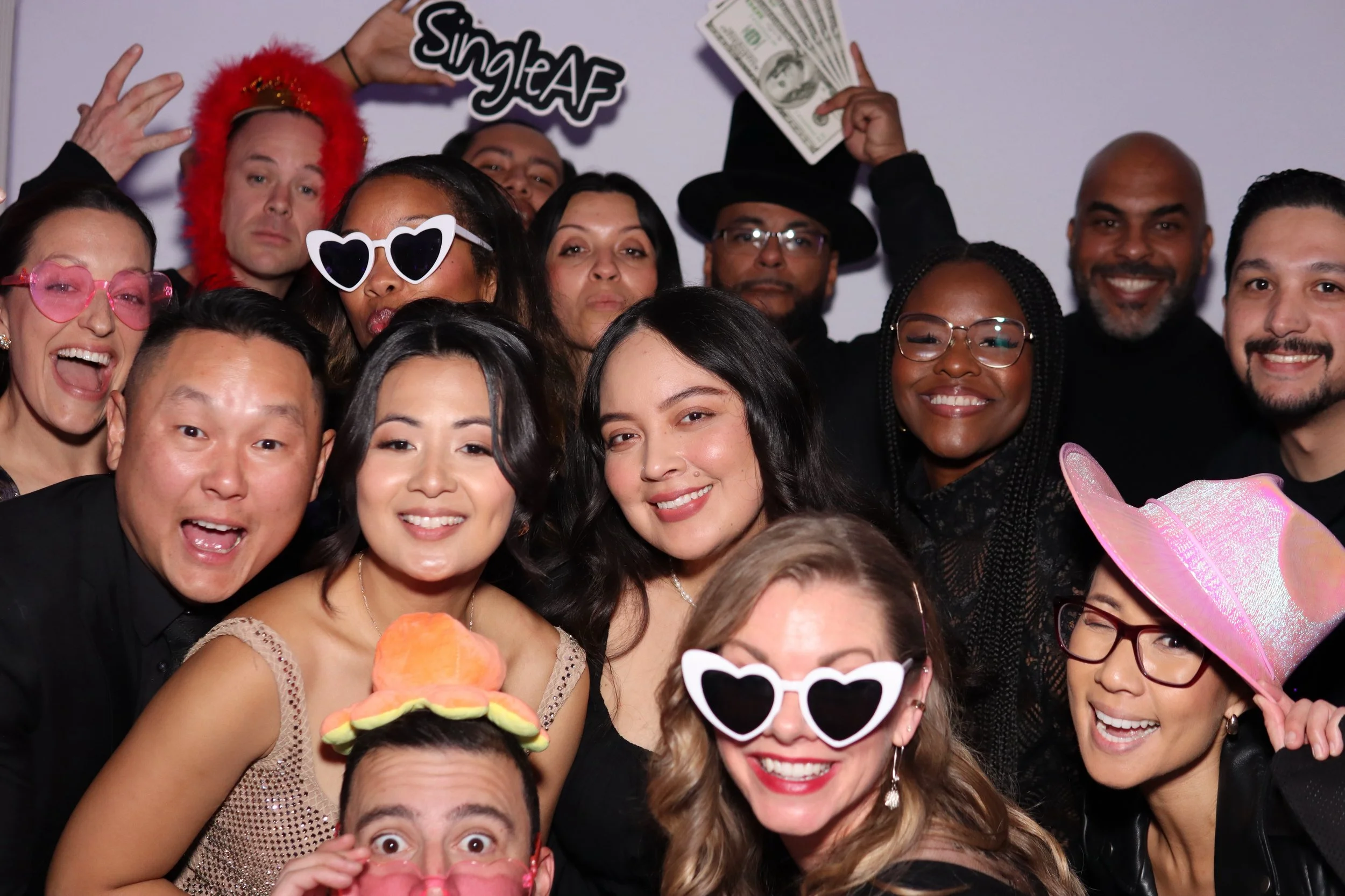 Group of people celebrating at a party, wearing fun accessories like oversized sunglasses, hats, and holding dollar bills, with a sign that says 'Single AF', smiling and posing for the camera.